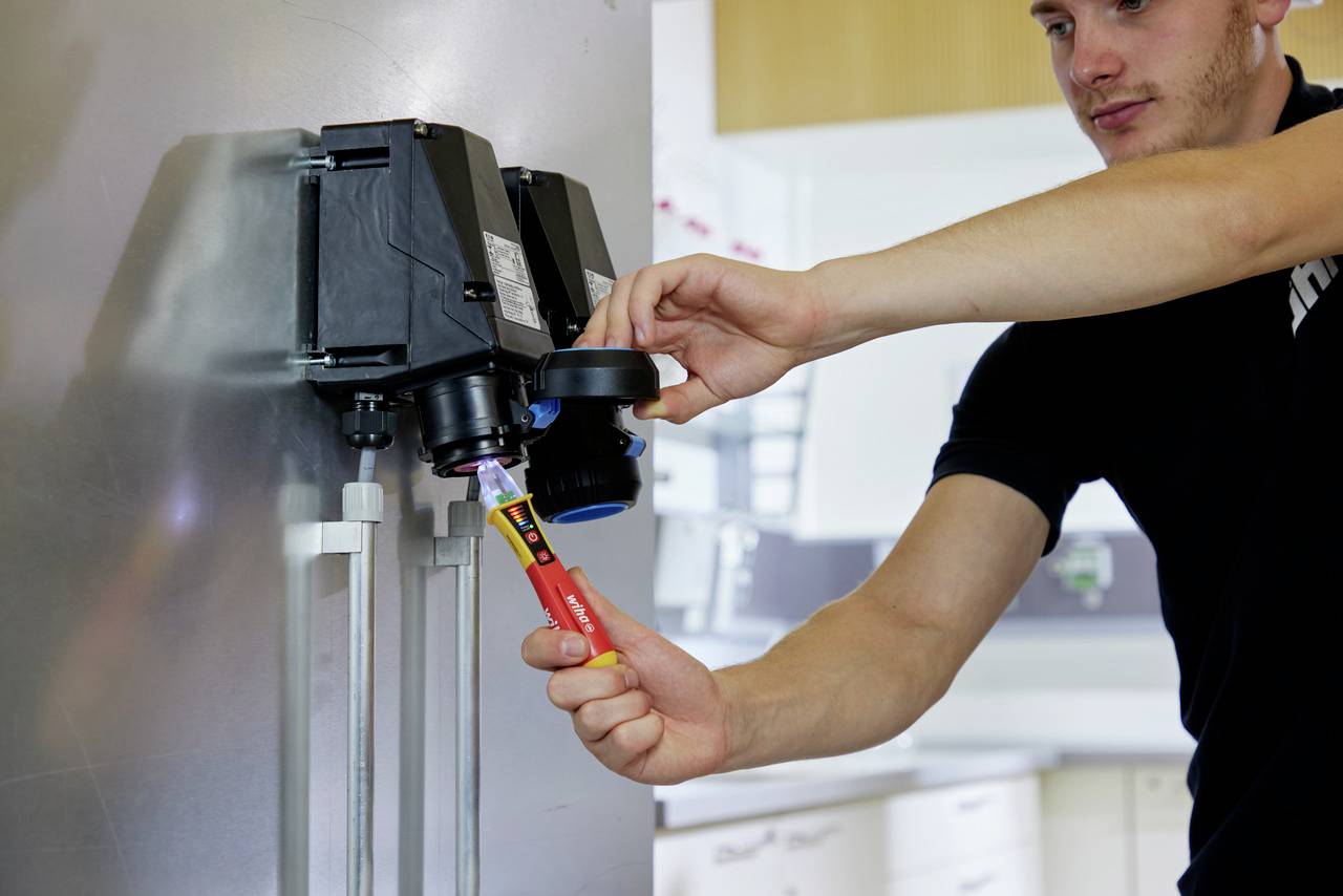 Muž kontroluje elektrickú súčiastku pomocou skúšačky napätia. Má na sebe čierne tričko a pracuje v modernej miestnosti.
