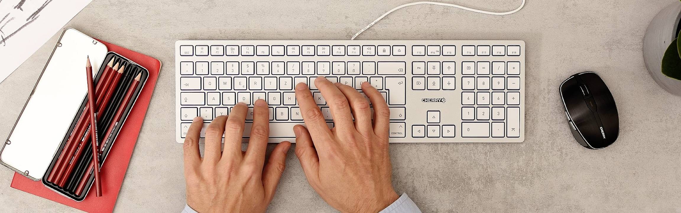 Osoba píše na bielej klávesnici vedľa červených ceruziek a čiernej myši na pracovnom stole.