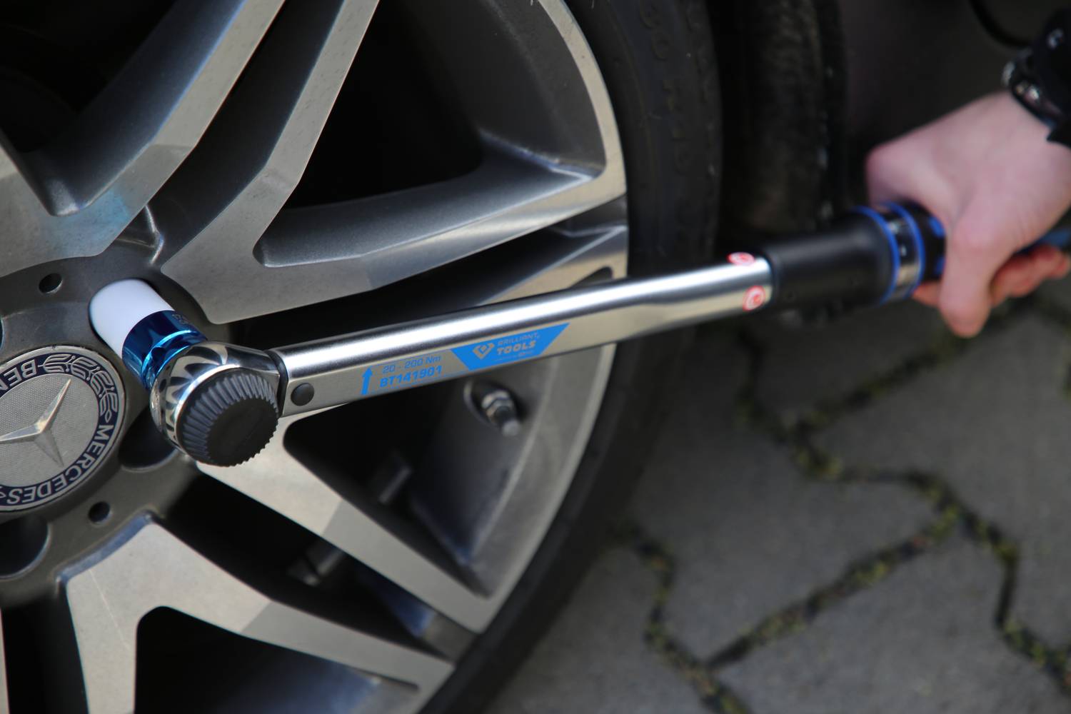 Jedna ruka používa momentový kľúč na dotiahnutie skrutiek automobilového kolesa. Koleso je strieborné s viacerými lúčmi.