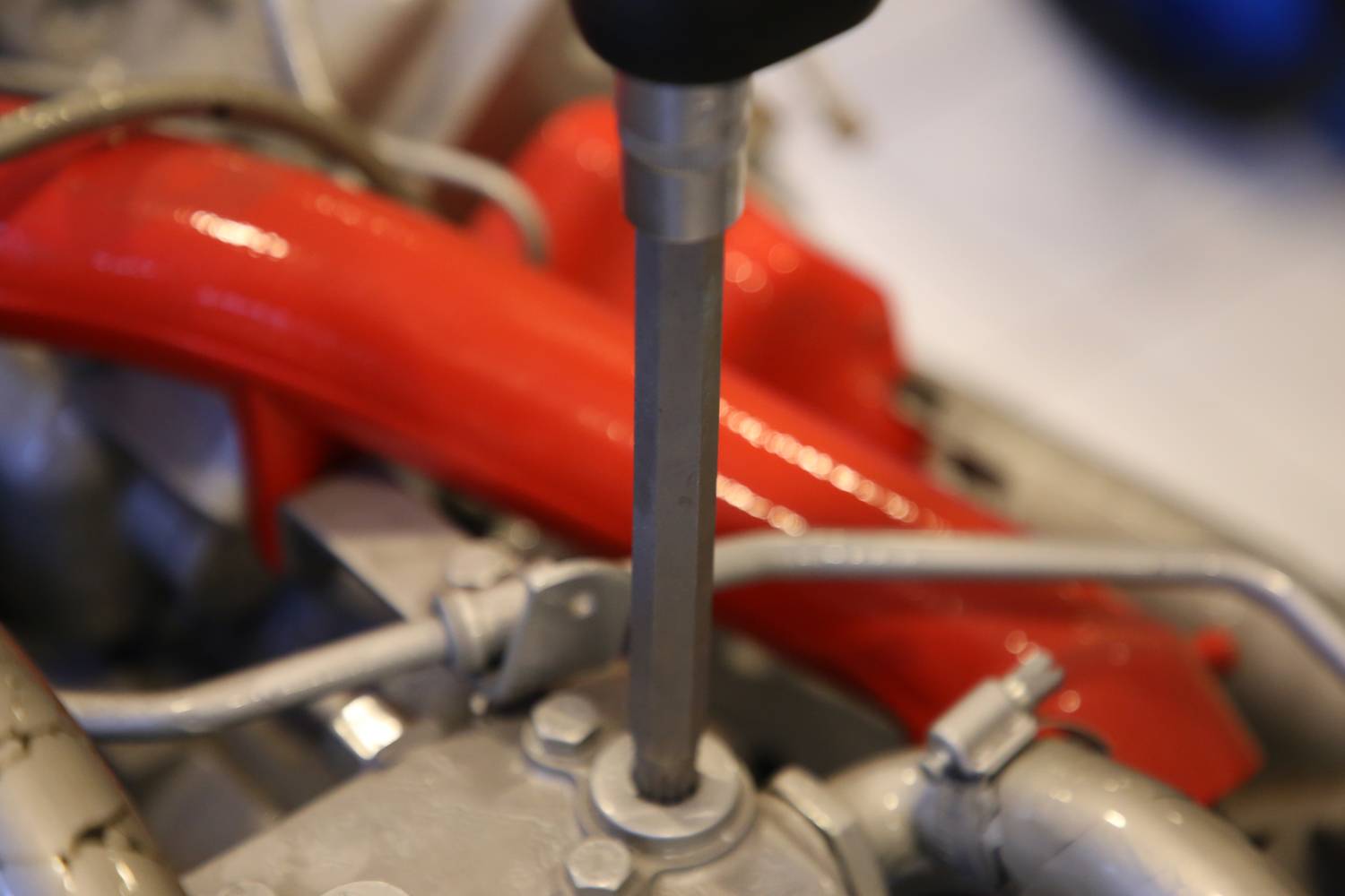Detailný záber na lesklú motorovú súčiastku s červenou rúrou a kovovými hadicami. V centre pozornosti je kľúč.