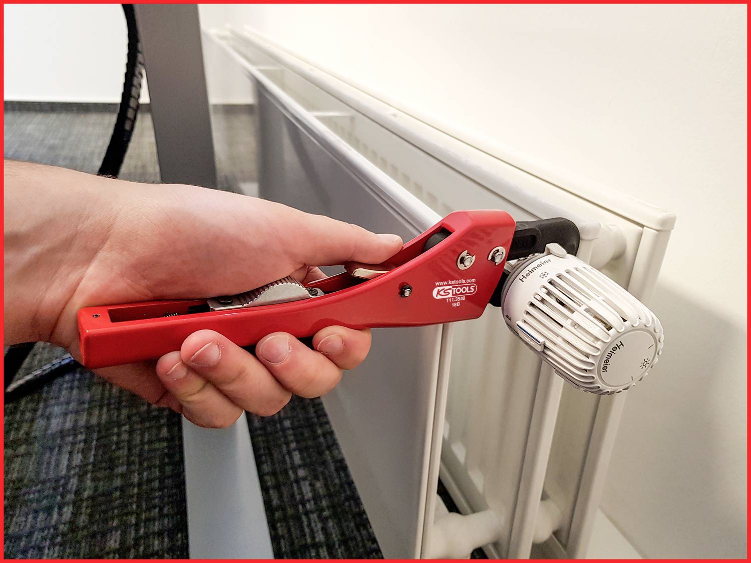 Jedna ruka drží červený nástroj a opatrne otáča termostatickým ventilom bieleho radiátora v kancelárii.