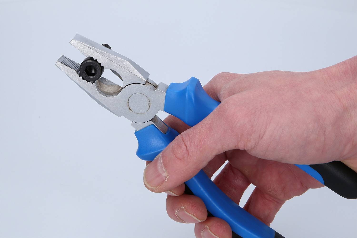 Ruka drží kliešte s modrým držadlom a striebornými čeľsťami, ktoré zachytávajú malé čierne ozubené koliesko.