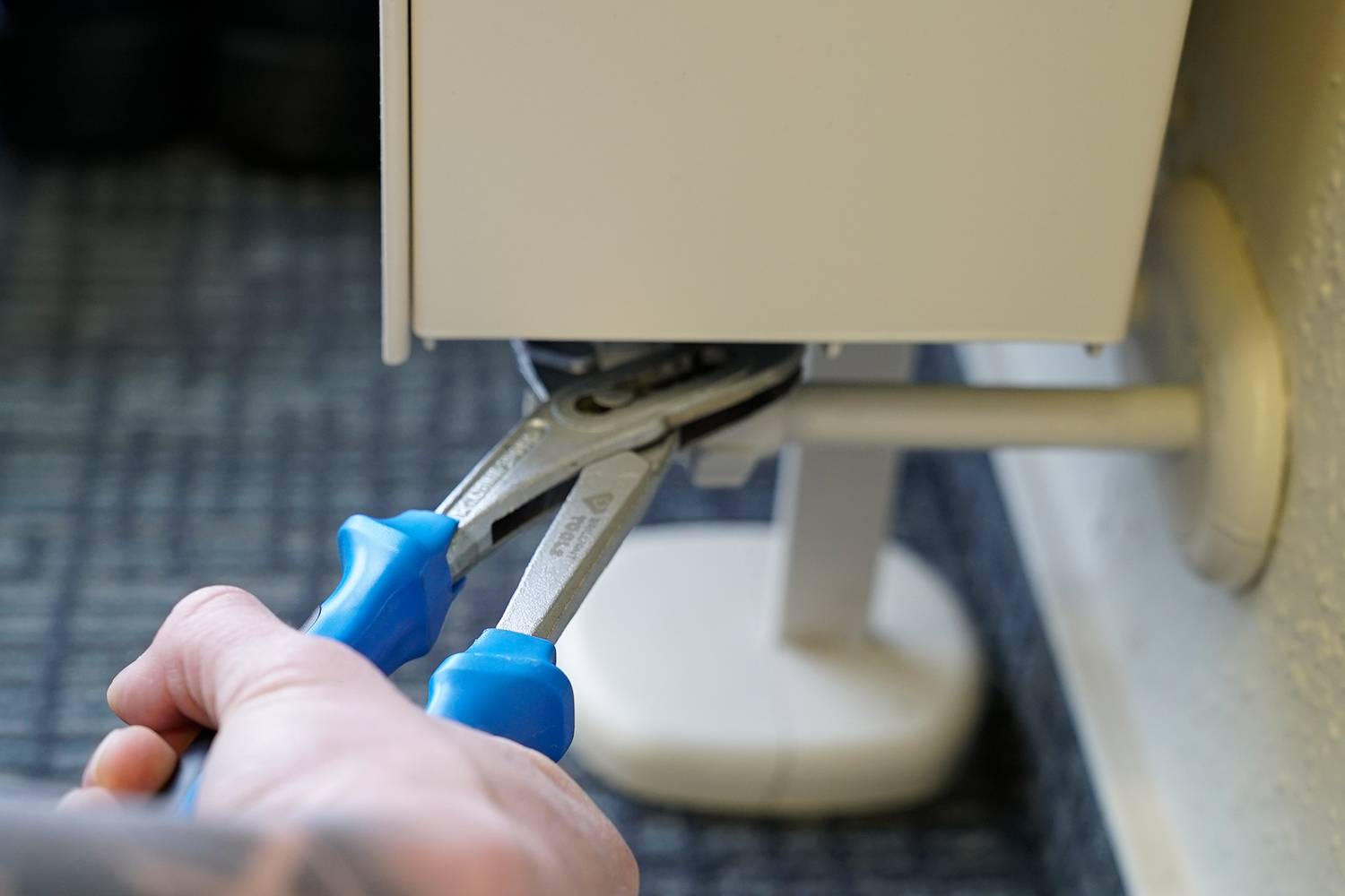 Ruka drží modré kliešte, ktoré pracujú na kovovej krabici pripevnenej na stene. Dôraz je kladený na manipuláciu s krabicou.