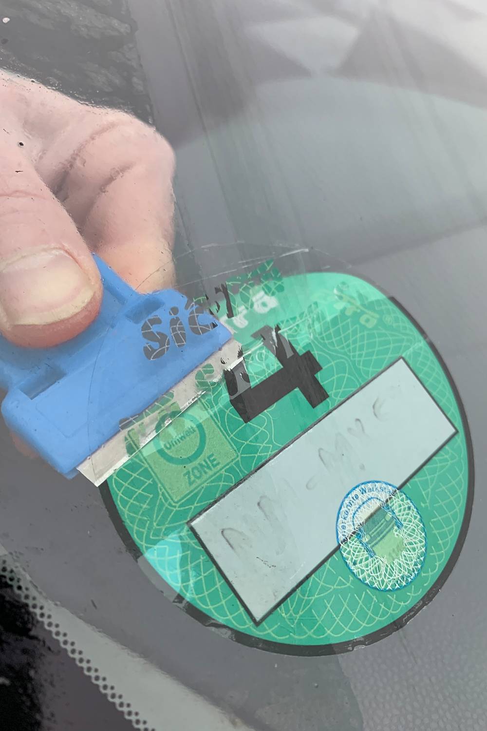 Ruka odstraňuje škrabkou zelenú nálepku emisnej kategórie 4 z čelného skla vozidla.