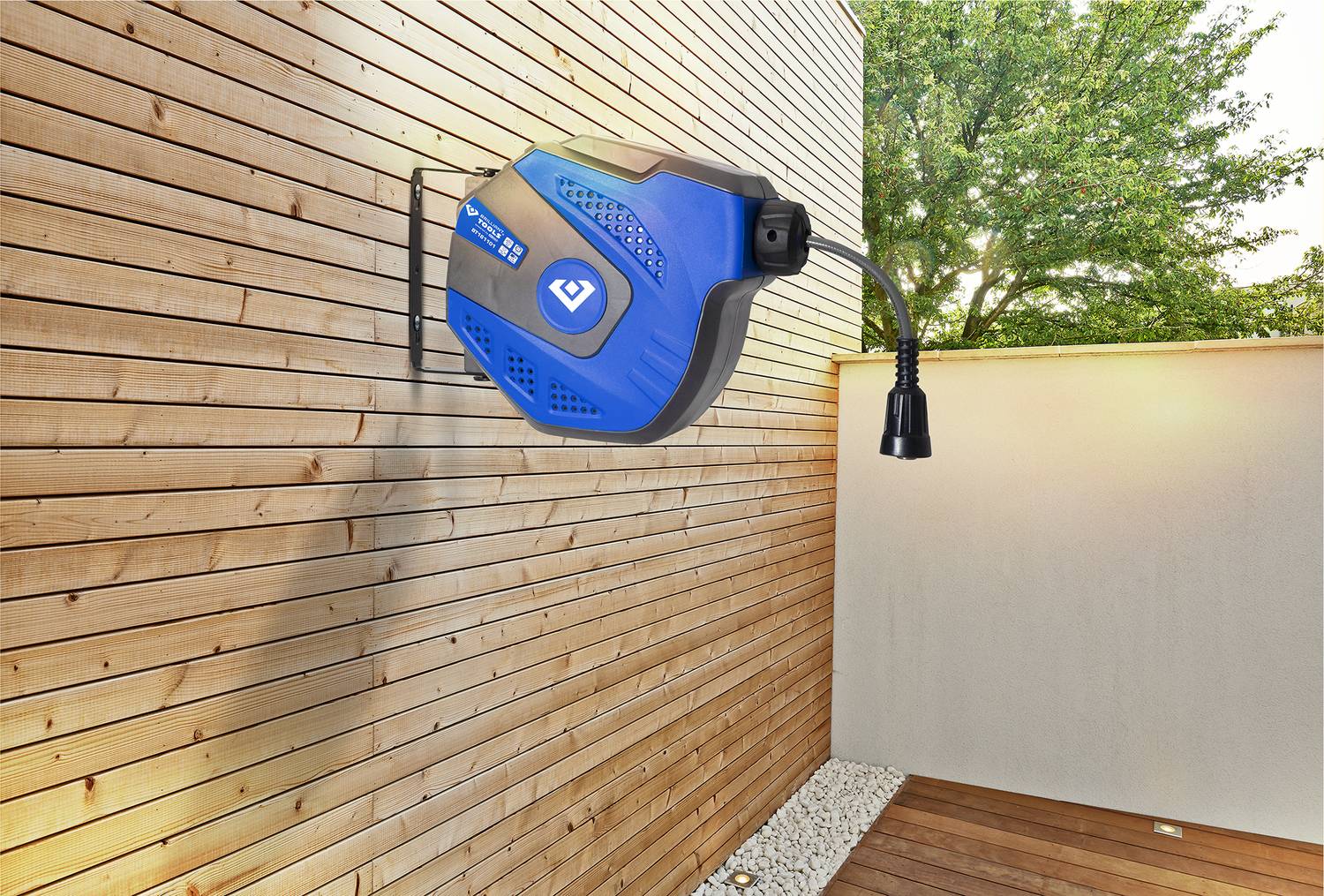 Nástenný modrý automatický navíjač záhradnej hadice namontovaný na drevenej fasáde, v pozadí so záhradou.