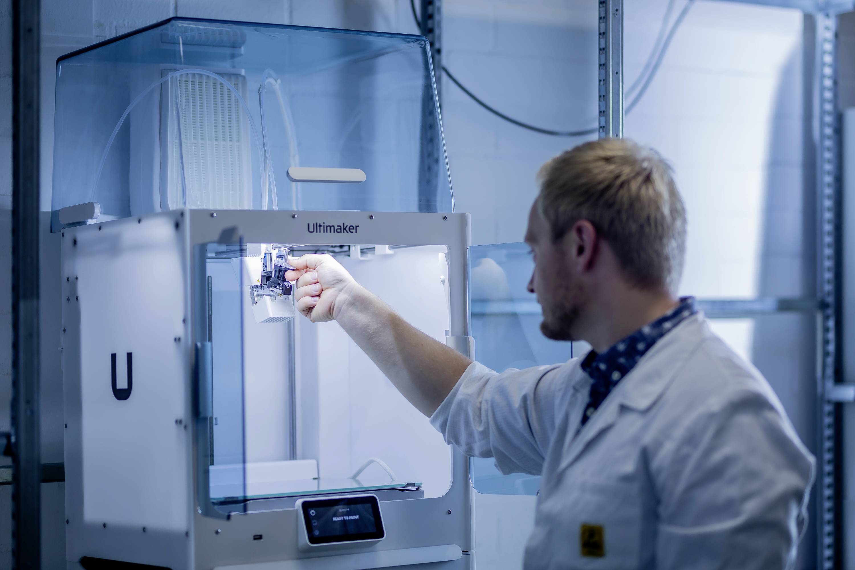 Muž v bielom laboratórnom plášti pracuje na 3D tlačiarni v laboratóriu. Kontroluje tlačiacu jednotku, počas jej prevádzky.