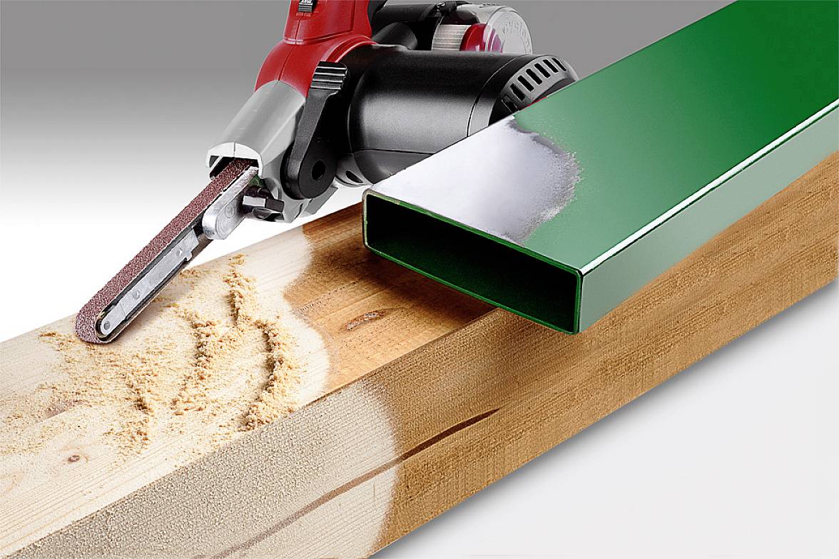 Elektrické brúsne zariadenie s červenou rukoväťou brúsi drevený trám, ktorý leží vedľa zelenej kovovej trubky.