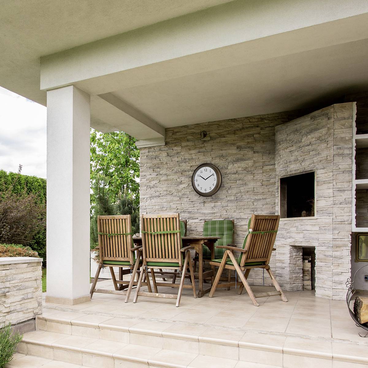 Terasa so stolom a stoličkami z dreva. Na stene visí hodiny. Vpravo je vidieť vstavaný gril.