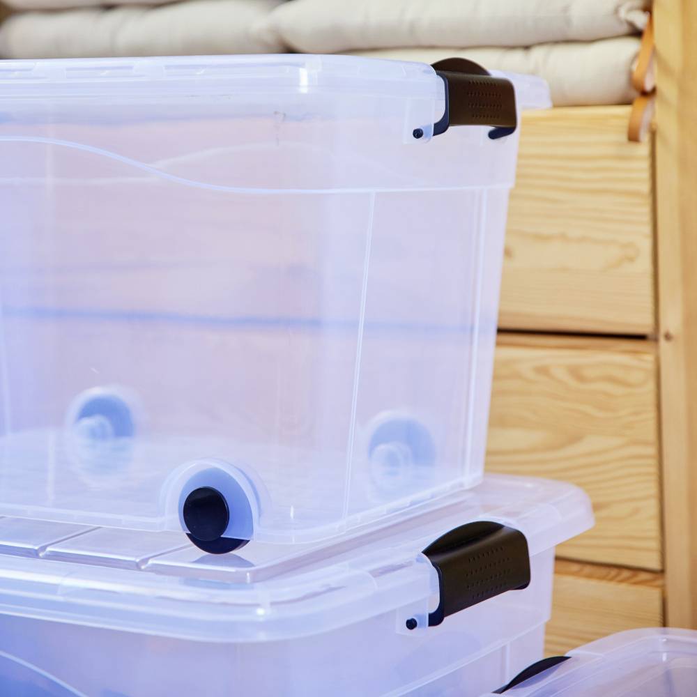 Priehľadné plastové boxy s čiernymi úchytkami sú naukladané na sebe. V pozadí sú viditeľné drevené nábytky. Ideálne na uskladnenie predmetov.