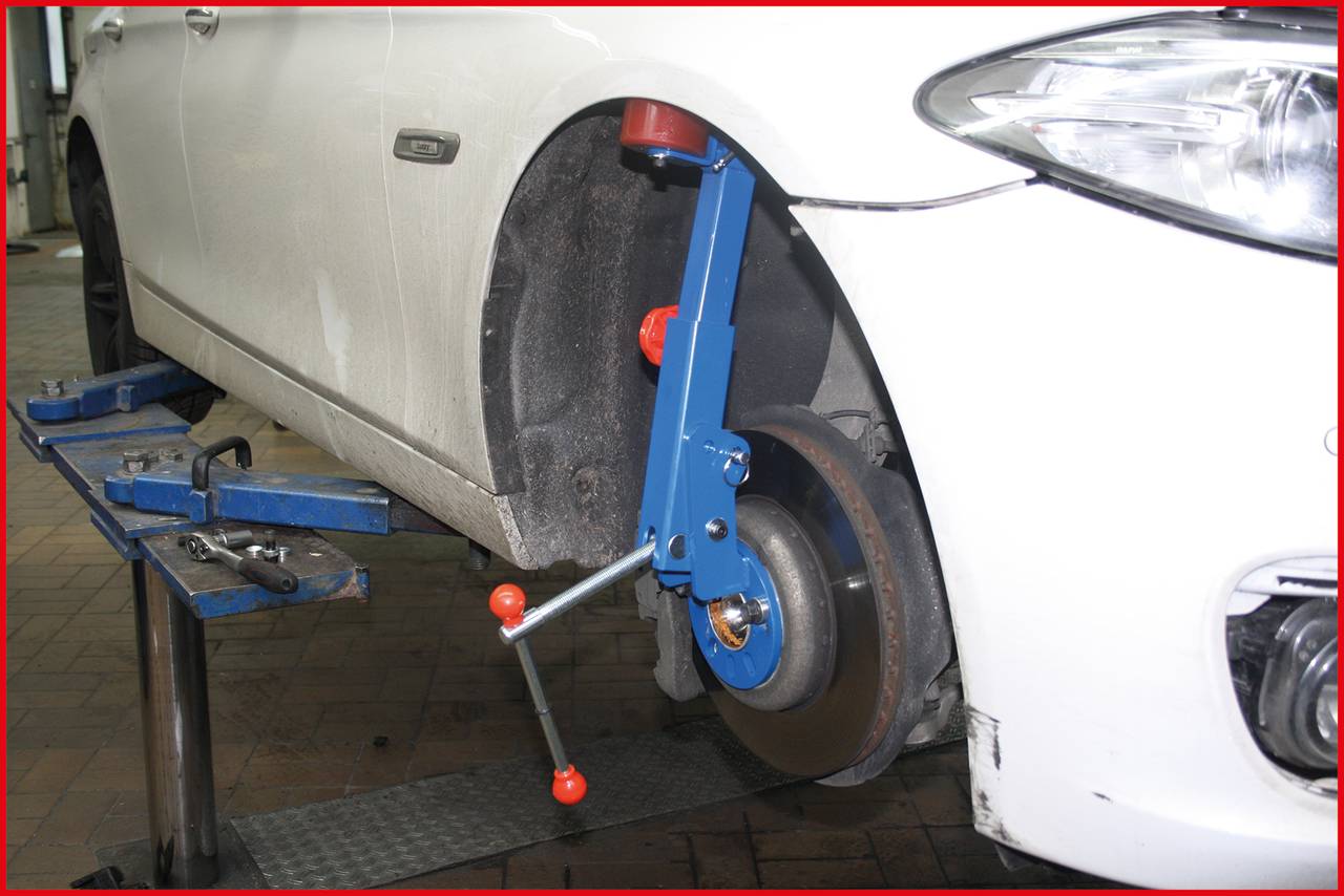 Biele auto v autoservise s kolesom vybaveným modrým zariadením na kontrolu geometrie nápravy.