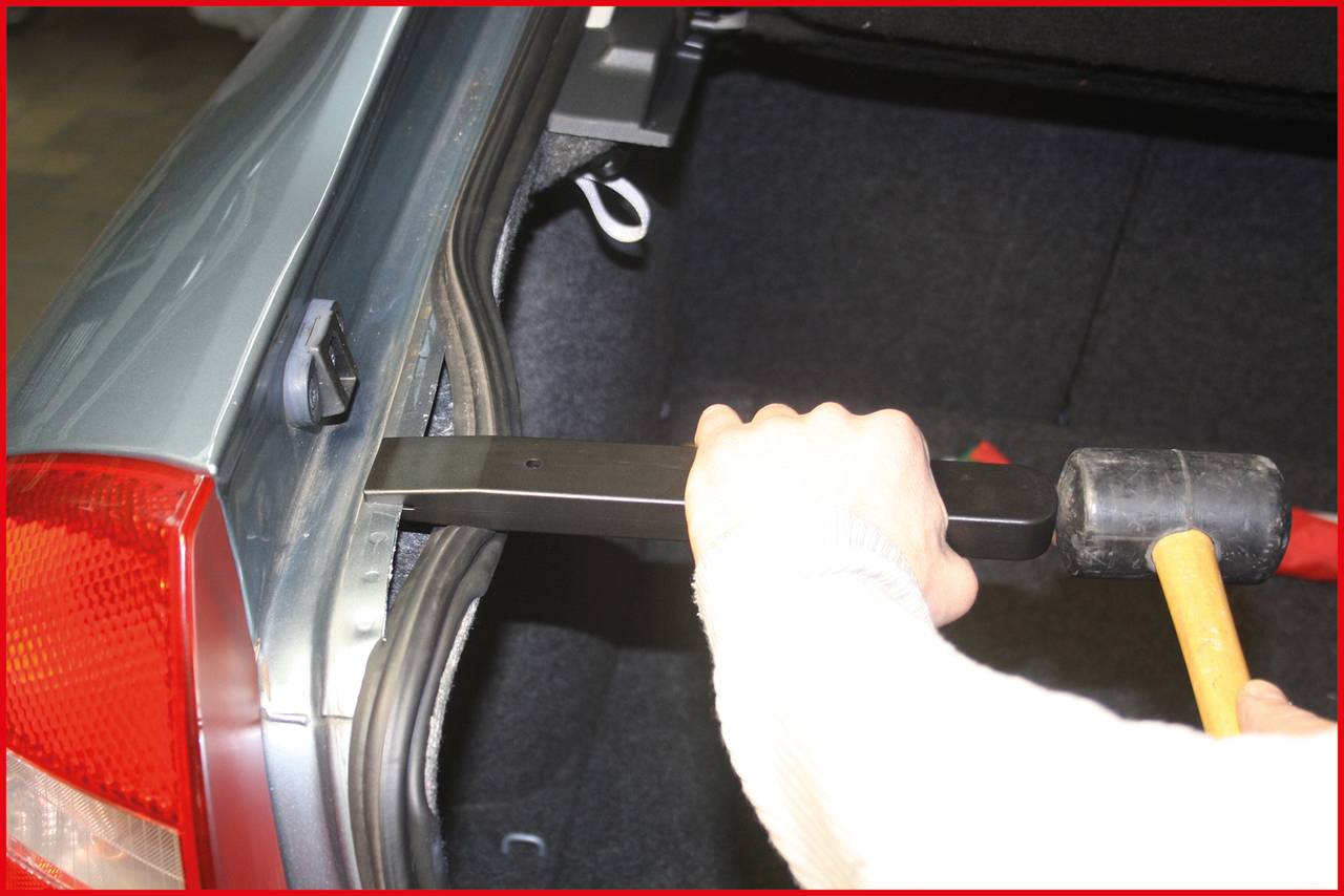 Osoba používa gumené kladivo, aby zatlačila dlhý nástroj do batožinového priestoru automobilu.
