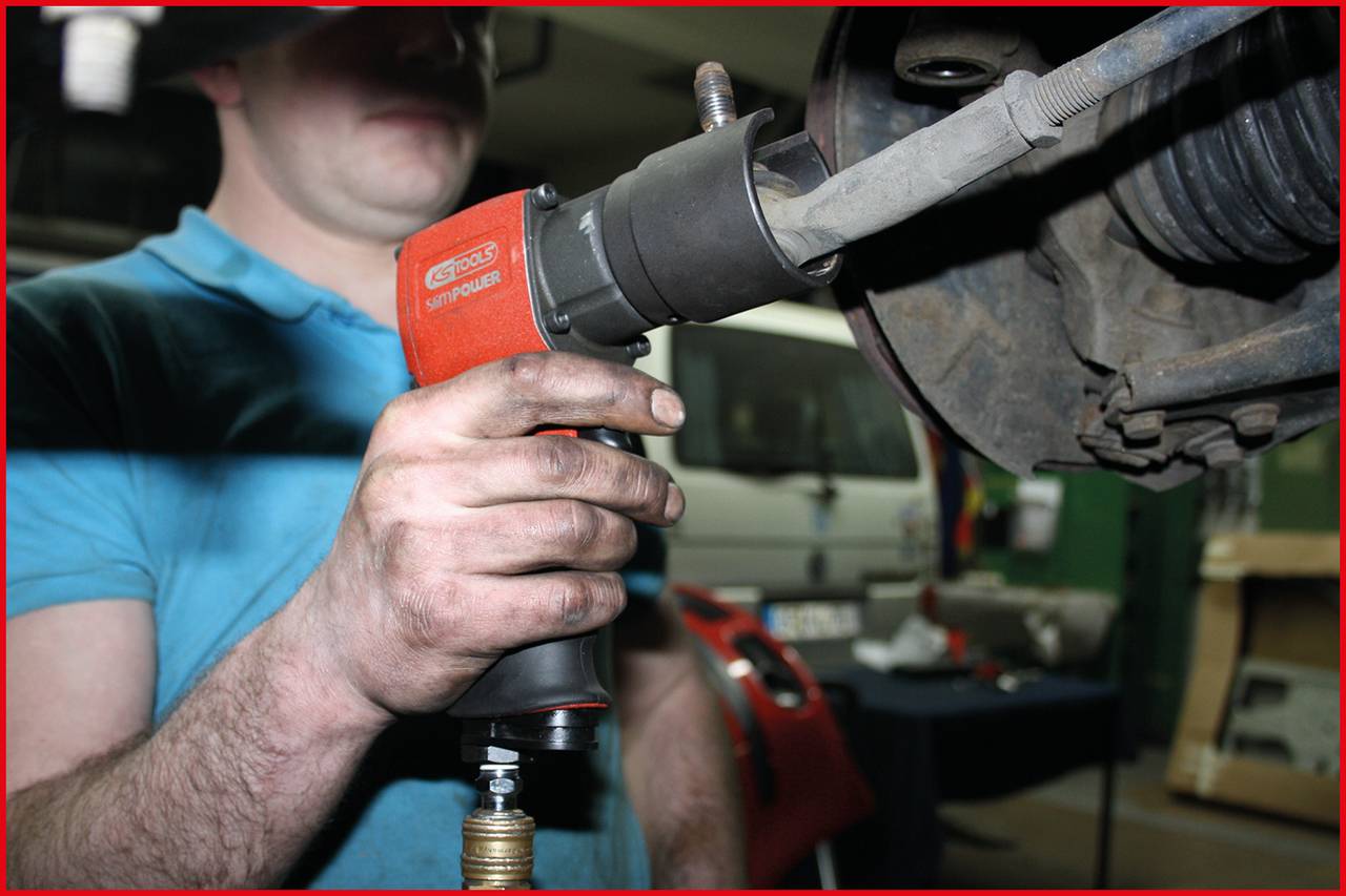 Mechanik používa pneumatický nástroj na práci na časti vozidla. V pozadí sú viditeľné nástroje a automobil.