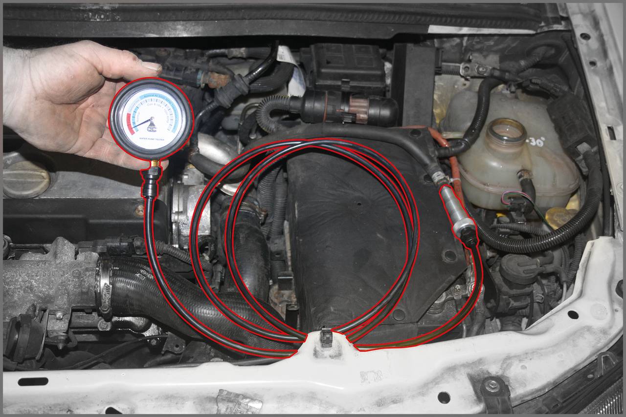 Jedna ruka drží manometer nad otvoreným motorovým priestorom automobilu. Červené hadice sú pripojené k motoru.