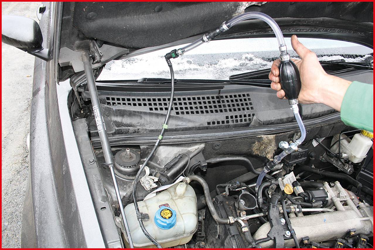 Ruka drží mechanickú pumpičku pripojenú k hadiciam v motorovom priestore automobilu. Motor je otvorený na účely údržby.