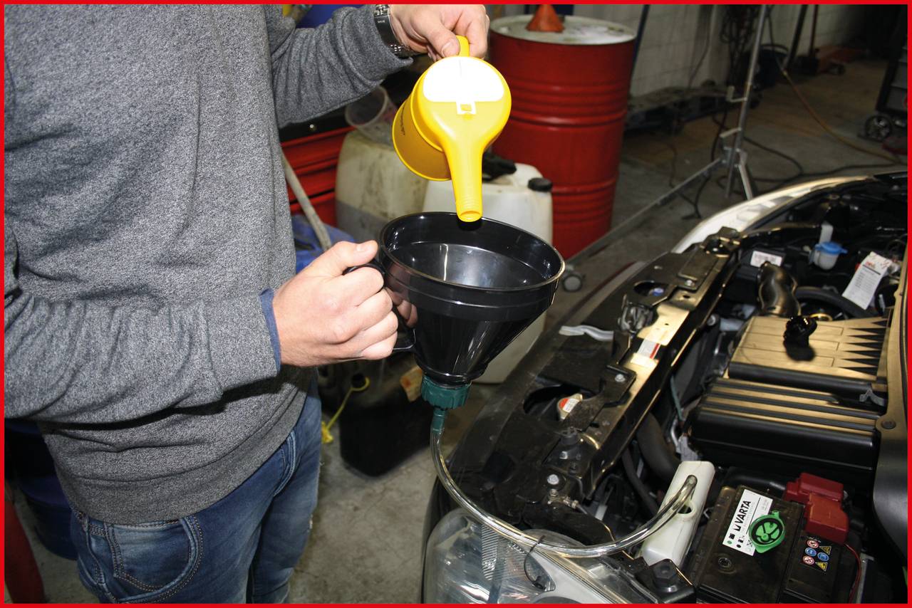 Osoba nalievaná motorový olej z žltého kanstra cez lievik do automobilu. V pozadí sú viditeľné sudové nádoby na olej.