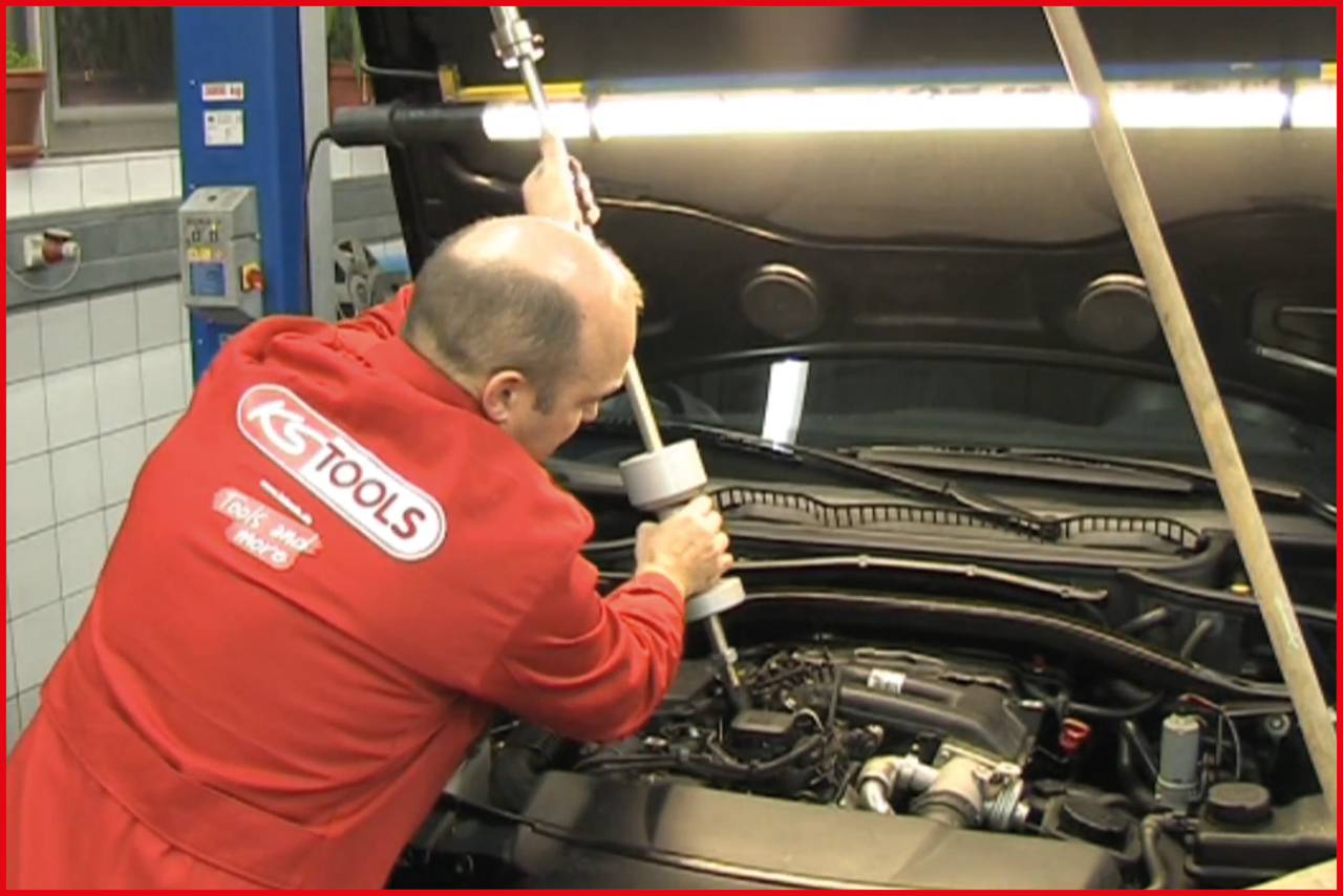 Mechanik v červenom pracovnom oblečení opravuje otvorený motorový priestor automobilu v dielni.