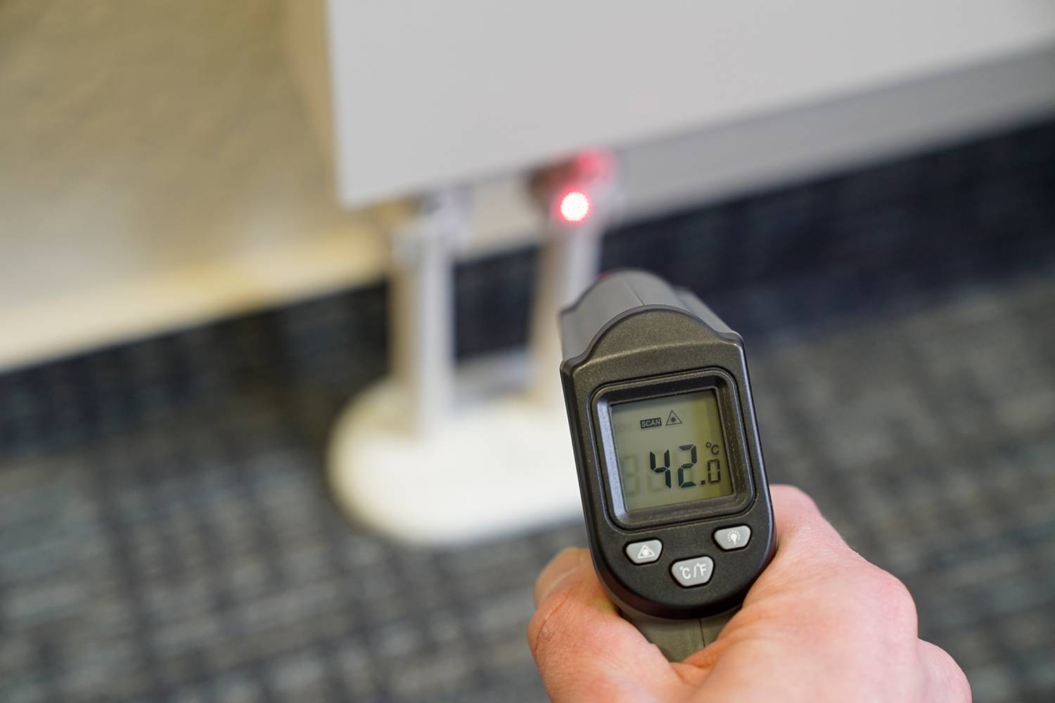 Osoba drží infračervený teplomer pred radiátorom, ktorý ukazuje 42,6 stupňov Celzia v momente merania.