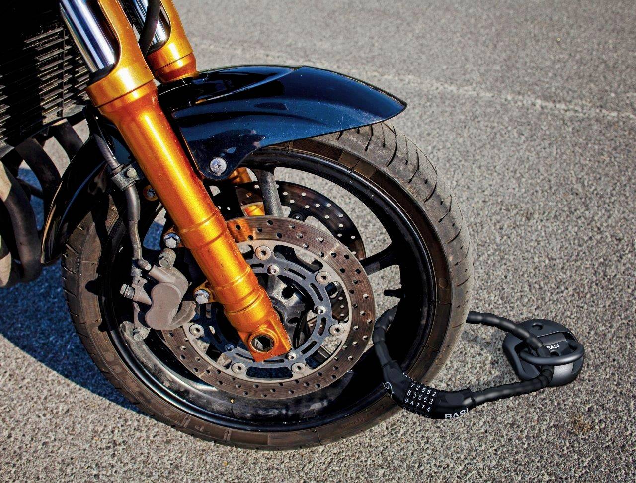 Predné motocyklové koleso s oranžovou vidlicou na asfaltovom povrchu, zabezpečené visacím zámkom na brzdovom kotúči.