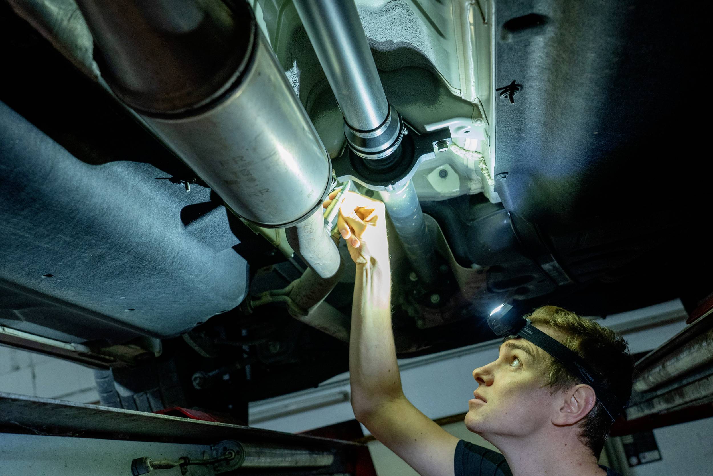 Mechanik kontroluje spodnú časť vozidla pomocou baterky v priestoroch dielne. Osvetlenie je zamerané na výfukový systém a elektroinštaláciu.
