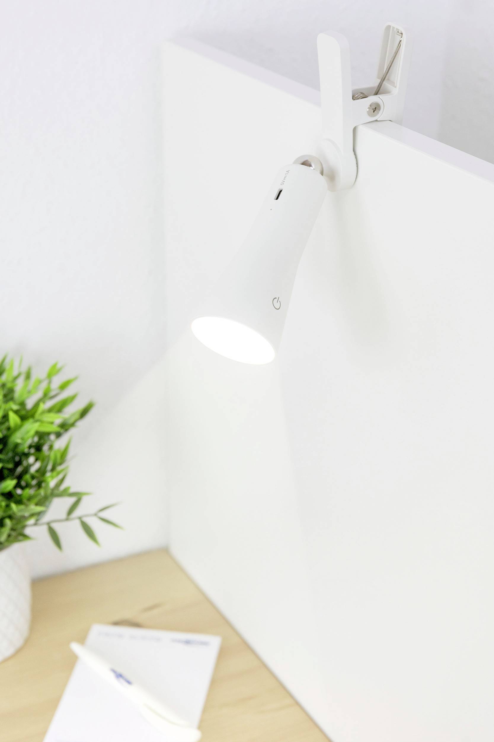 Biela štipková lampa osvetľuje roh písacieho stola, vedľa poznámkového bloku a pera. Vľavo je kvetináč s rastlinou.