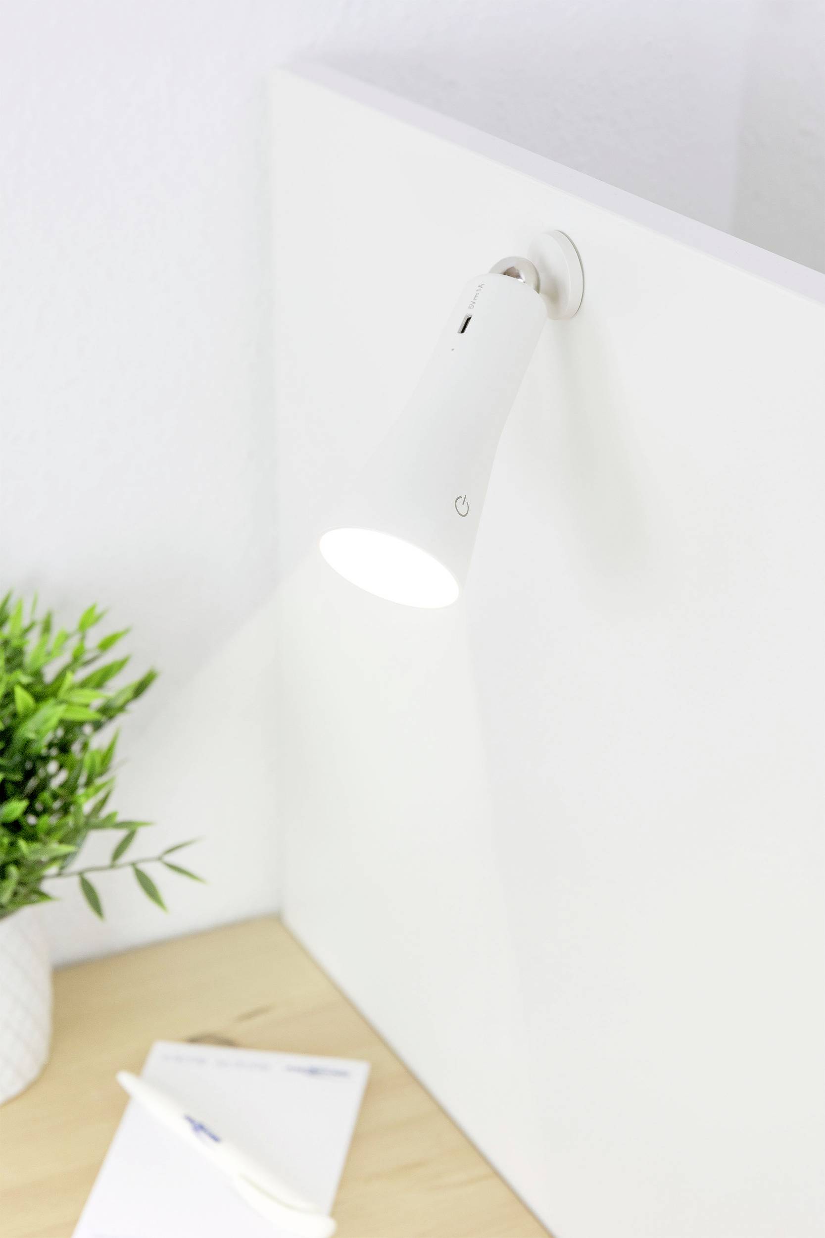 Lampa je upevnená na bielej stene a svieti. Vedľa nej stojí rastlina v kvetináči a na stole leží papier s perom.