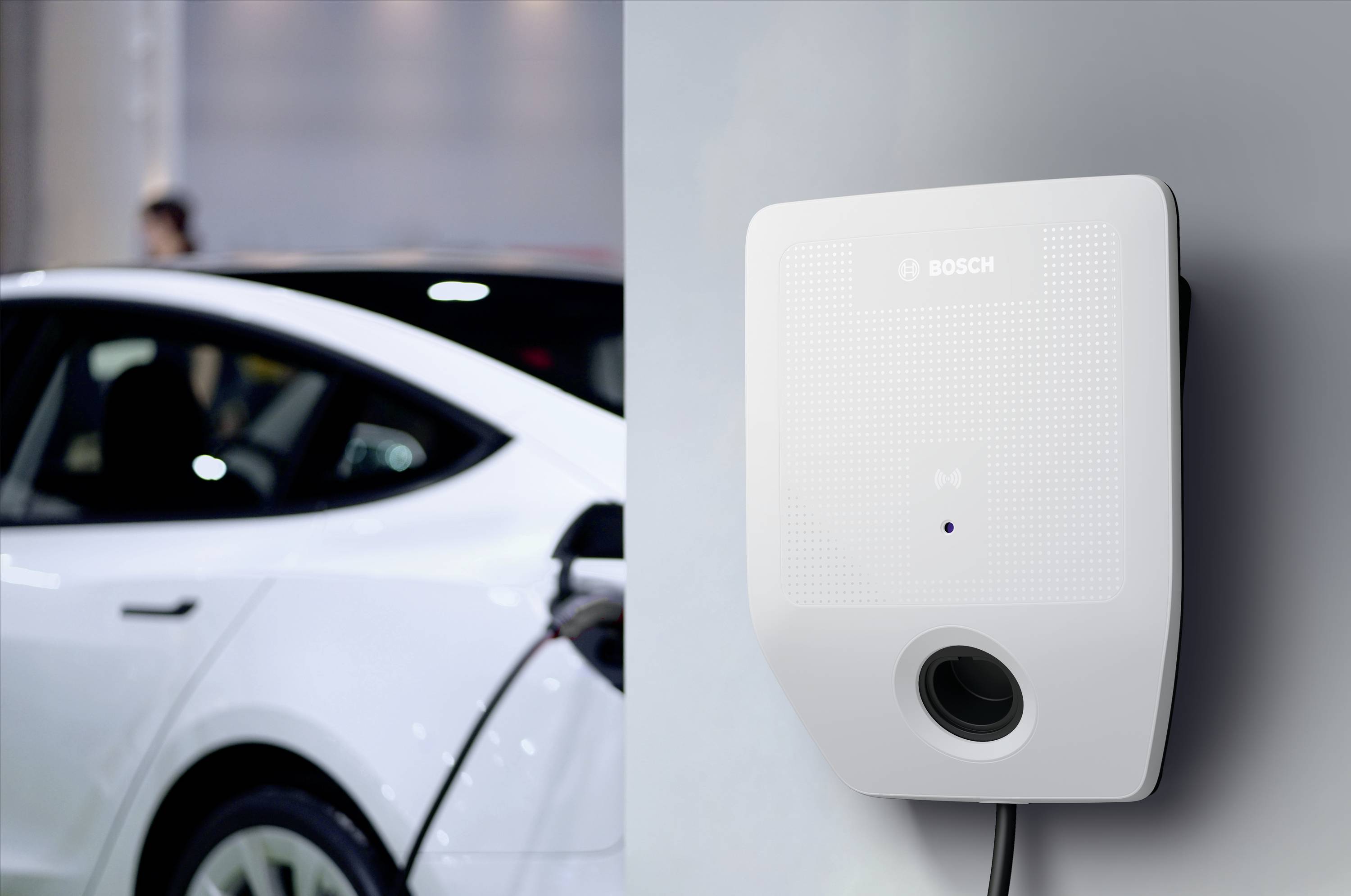 Elektrické auto sa nabíja na bielej nabíjacej stanici. Nabíjacia stanica je namontovaná na stene a auto je čiastočne viditeľné.