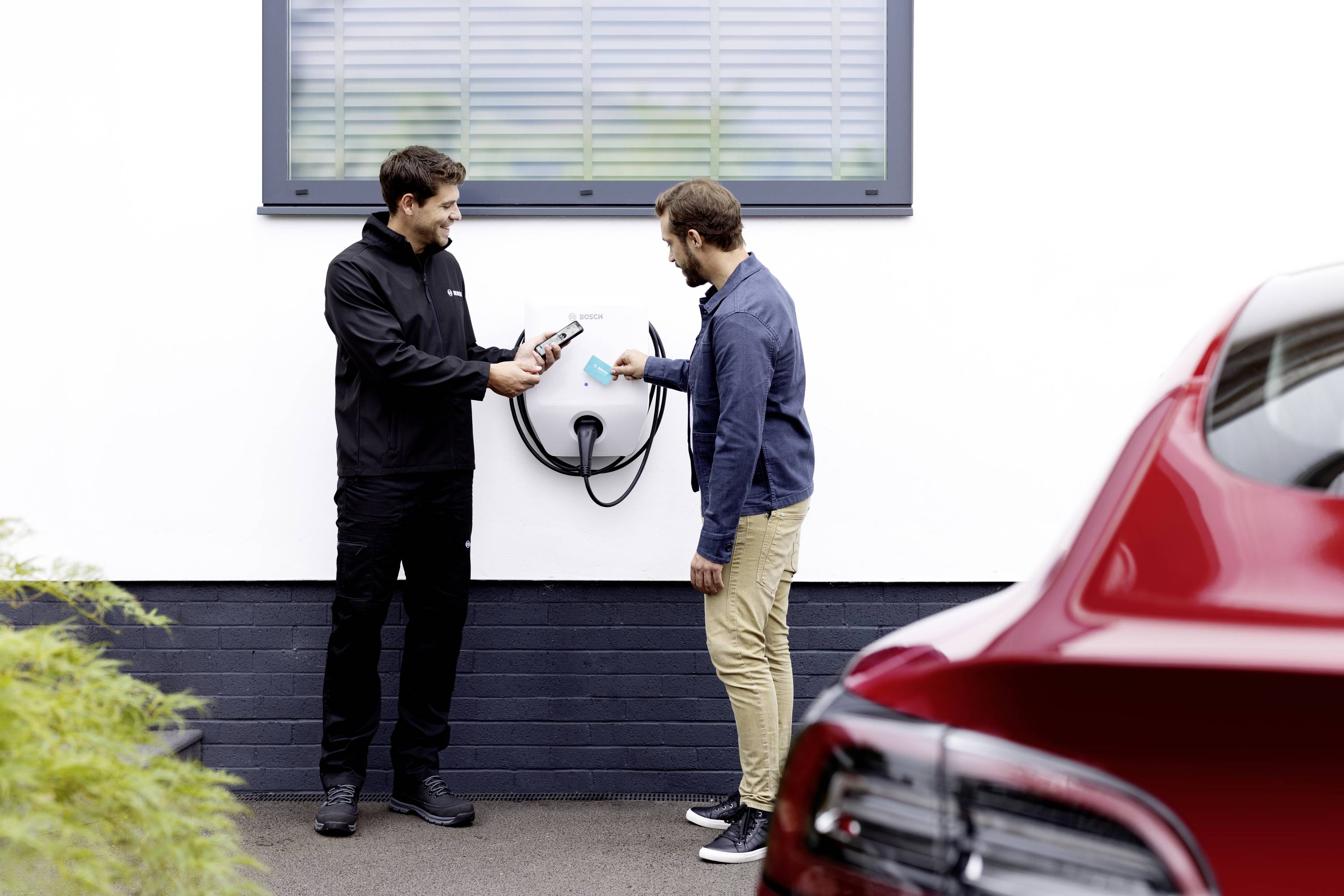 Dvaja muži stoja pred bielou stenou vedľa nabíjacej stanice pre elektromobily. Jeden drží smartfón, druhý kábel. V popredí je viditeľné červené auto.