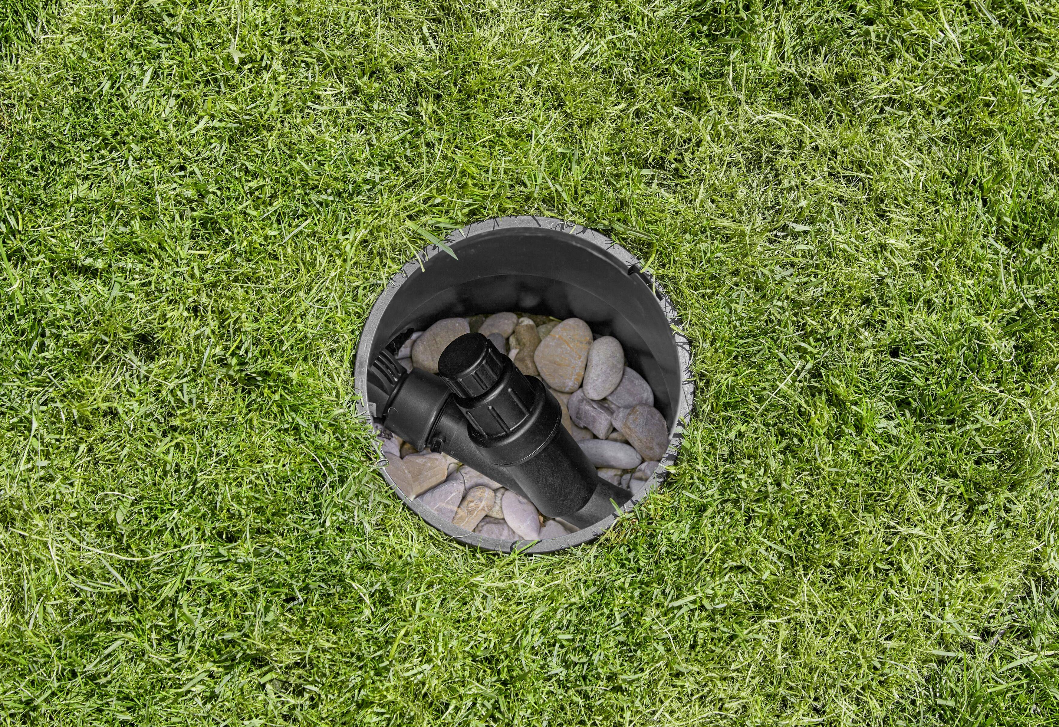 Čierny zavlažovací hlavicový postrekovač je obklopený kruhovým otvorom na trávnatej ploche, s kameňmi vo vnútri tohto otvoru.