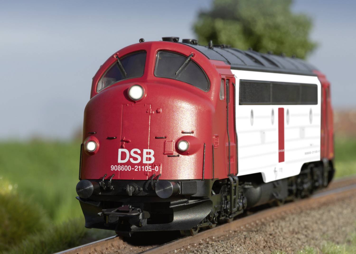 Červený vlak DSB jazdí po koľaji vo vidieckom prostredí. Na prednej strane vlaku je vyznačené číslo '906800-21105-01'.
