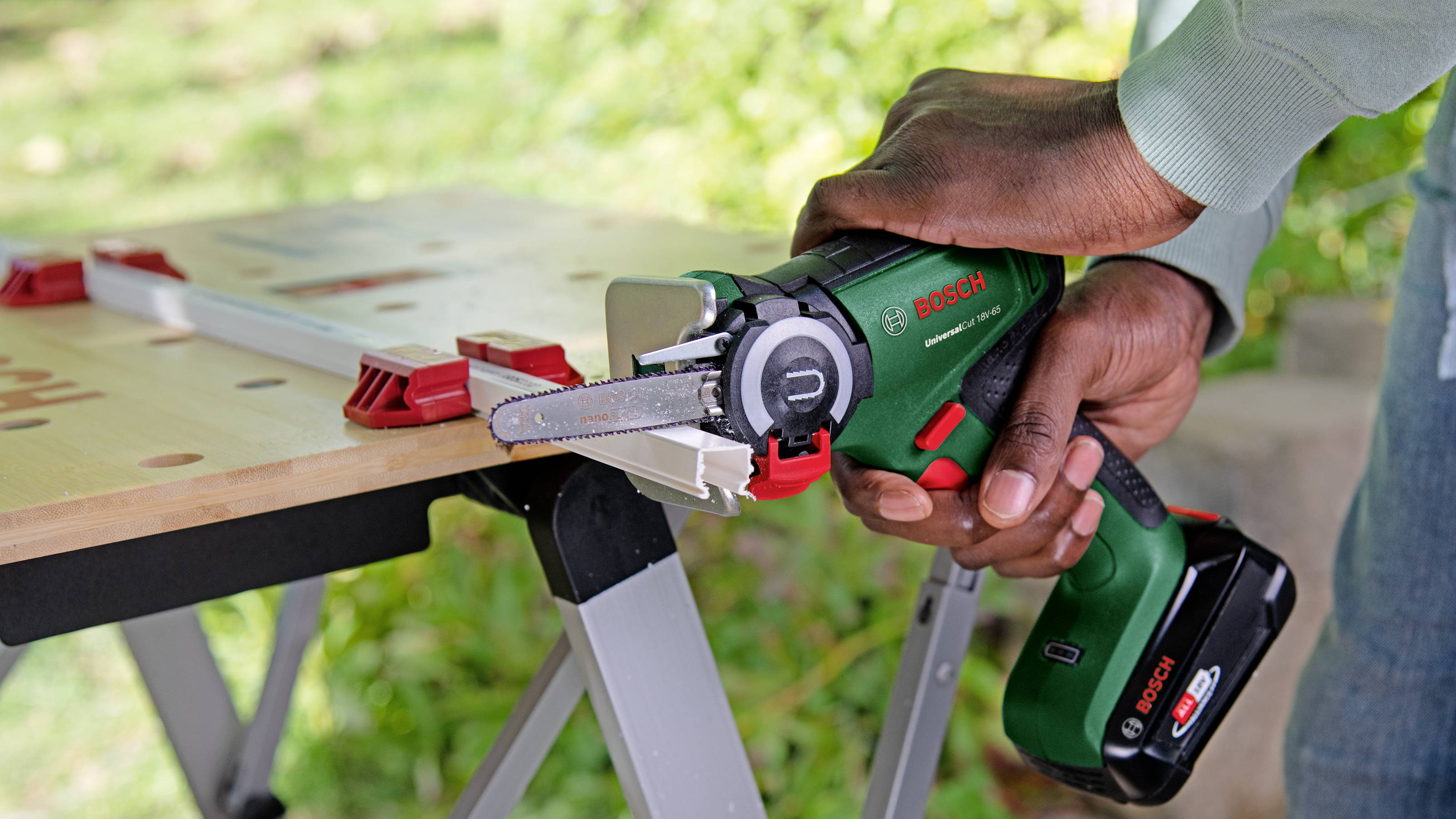 Osoba používa zelenú bezdrôtovú mini-reťazovú pílu na pracovnom stole vonku.