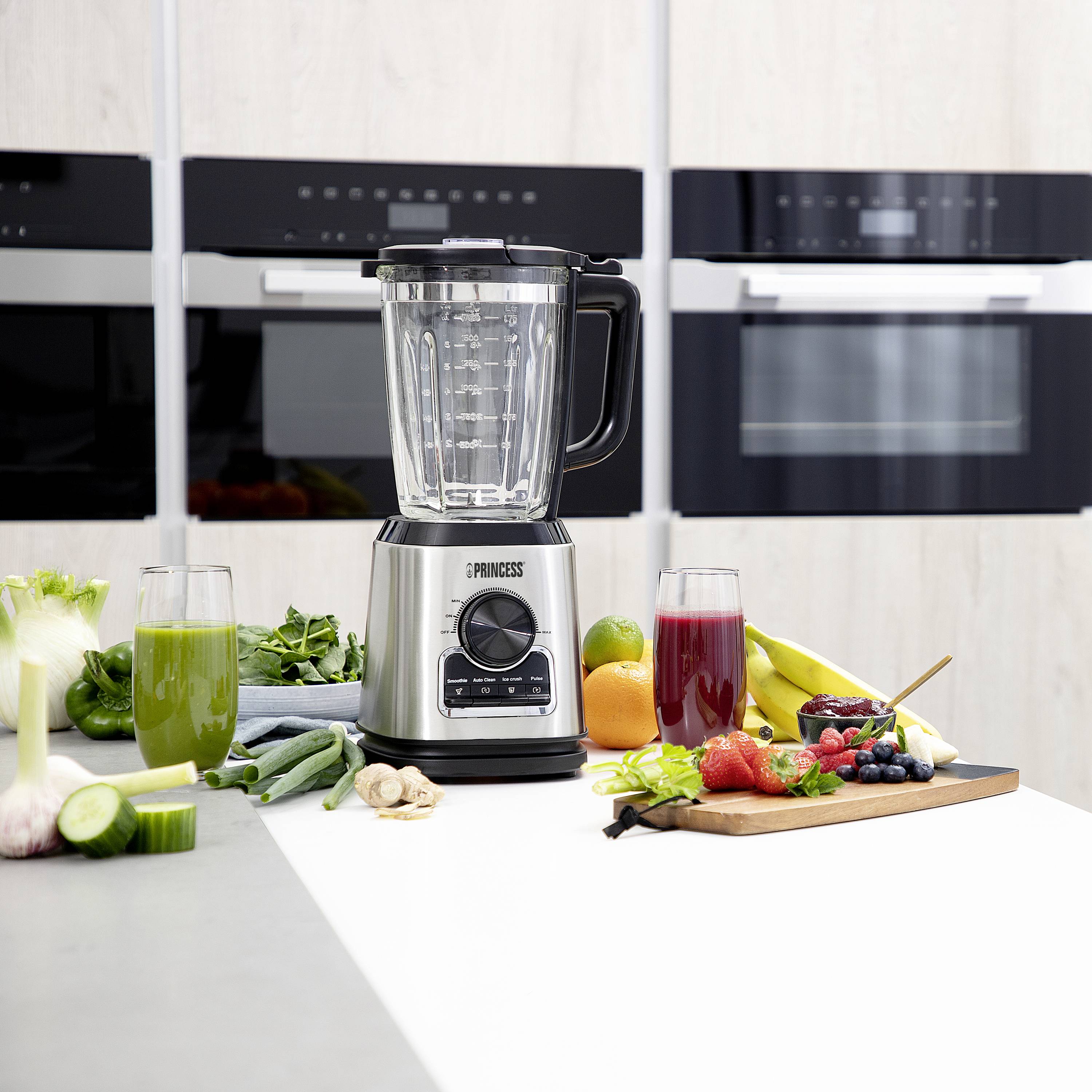 Mixér na kuchynskej linke, obklopený čerstvou zeleninou, ovocím a dvoma pohármi smoothies v zelenej a červenej farbe. V pozadí kuchynské spotrebiče.