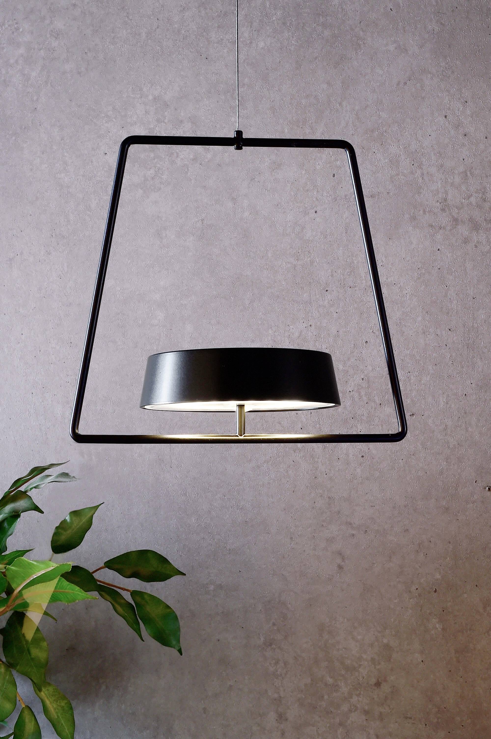 Moderná závesná lampa v geometrickom dizajne pred sivou stenou. V ľavom dolnom rohu sú viditeľné zelené listy rastliny.