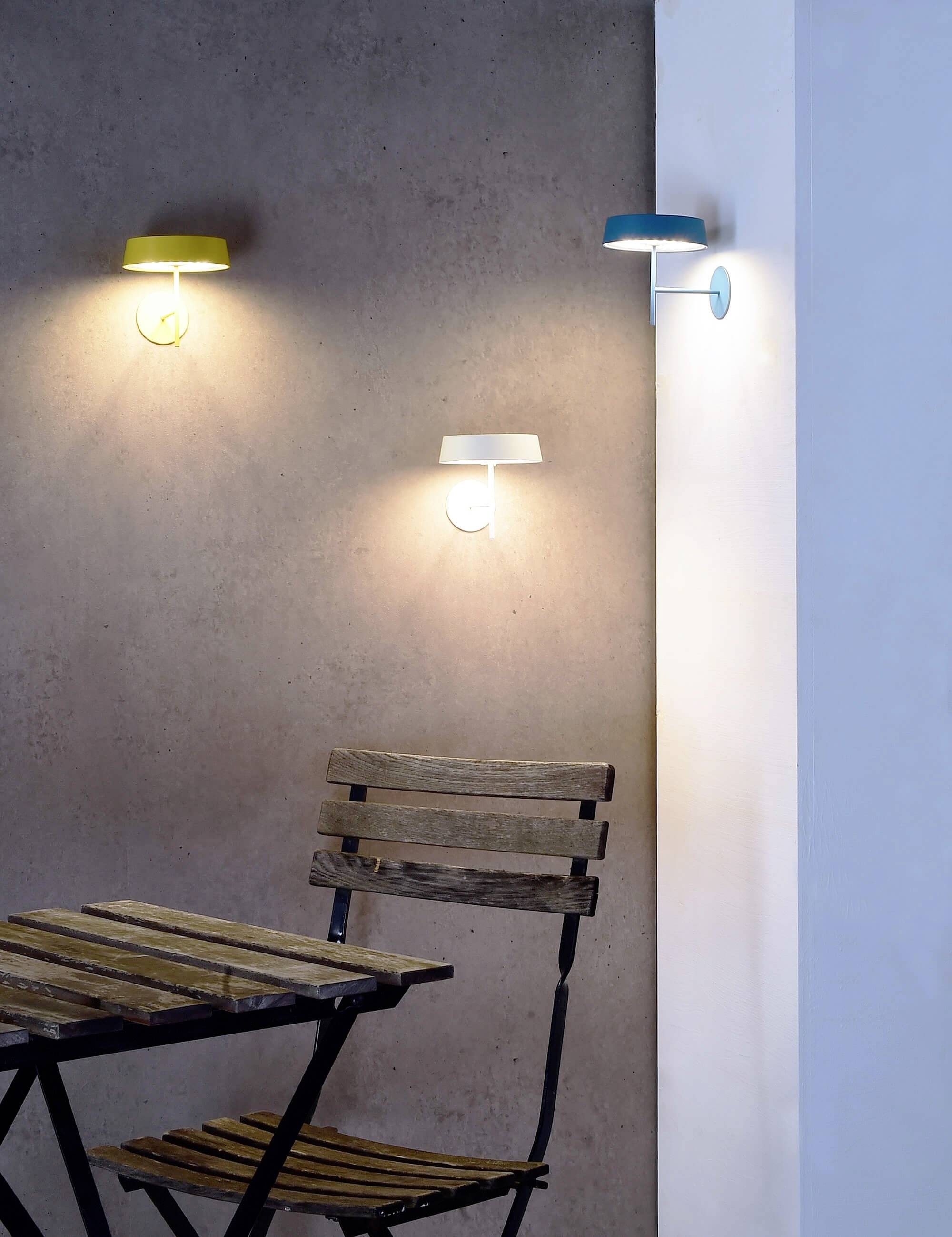 Drevená zostava stola a stoličiek stojí pred stenou s tromi modernými, rôznofarebne prevedenými nastennými lampami, ktoré svietia.