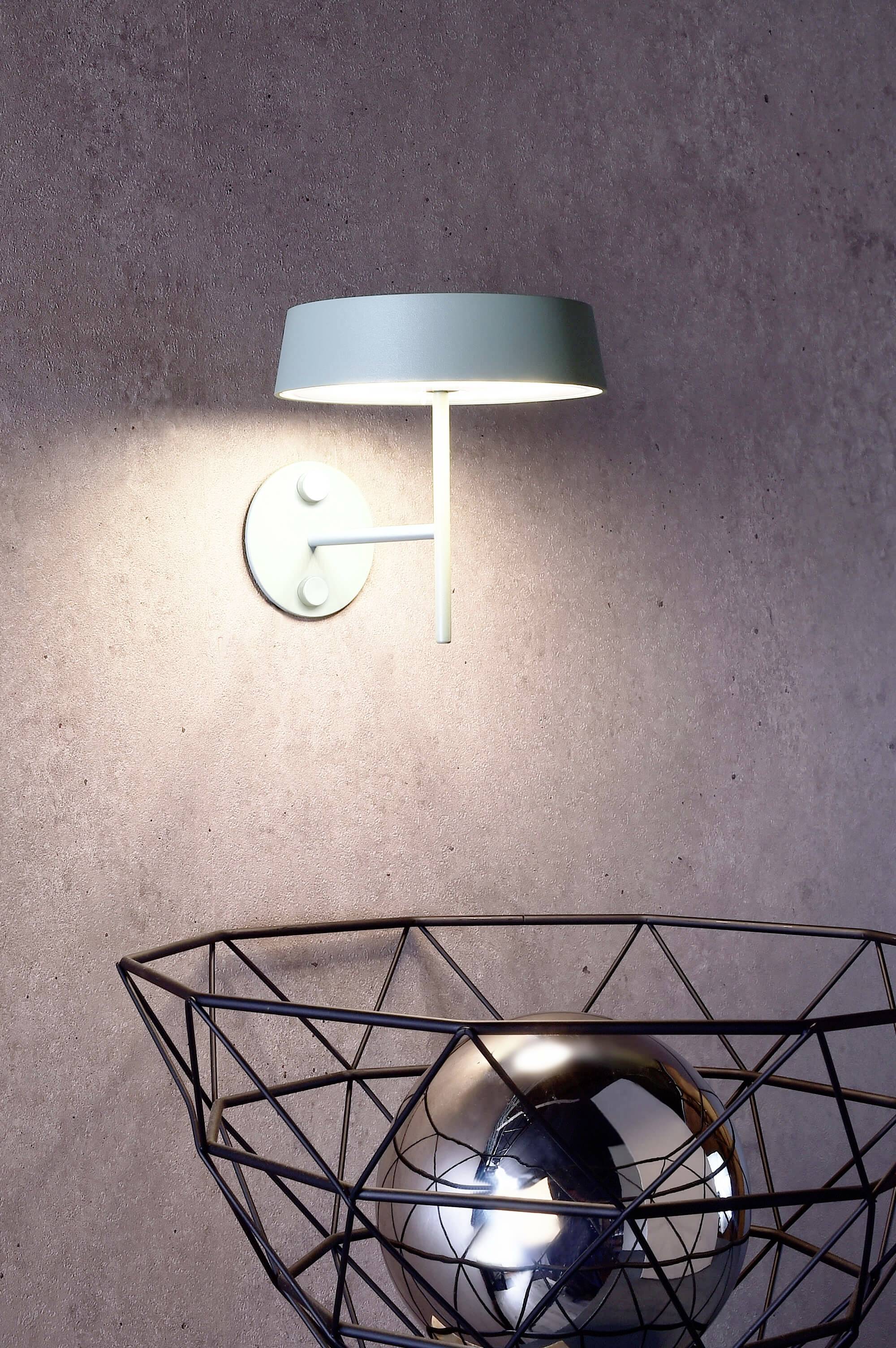 Moderná nástenná lampa vyžaruje svetlo na kovovú misu s lesklým guľovým objektom na textúrovanej stene.