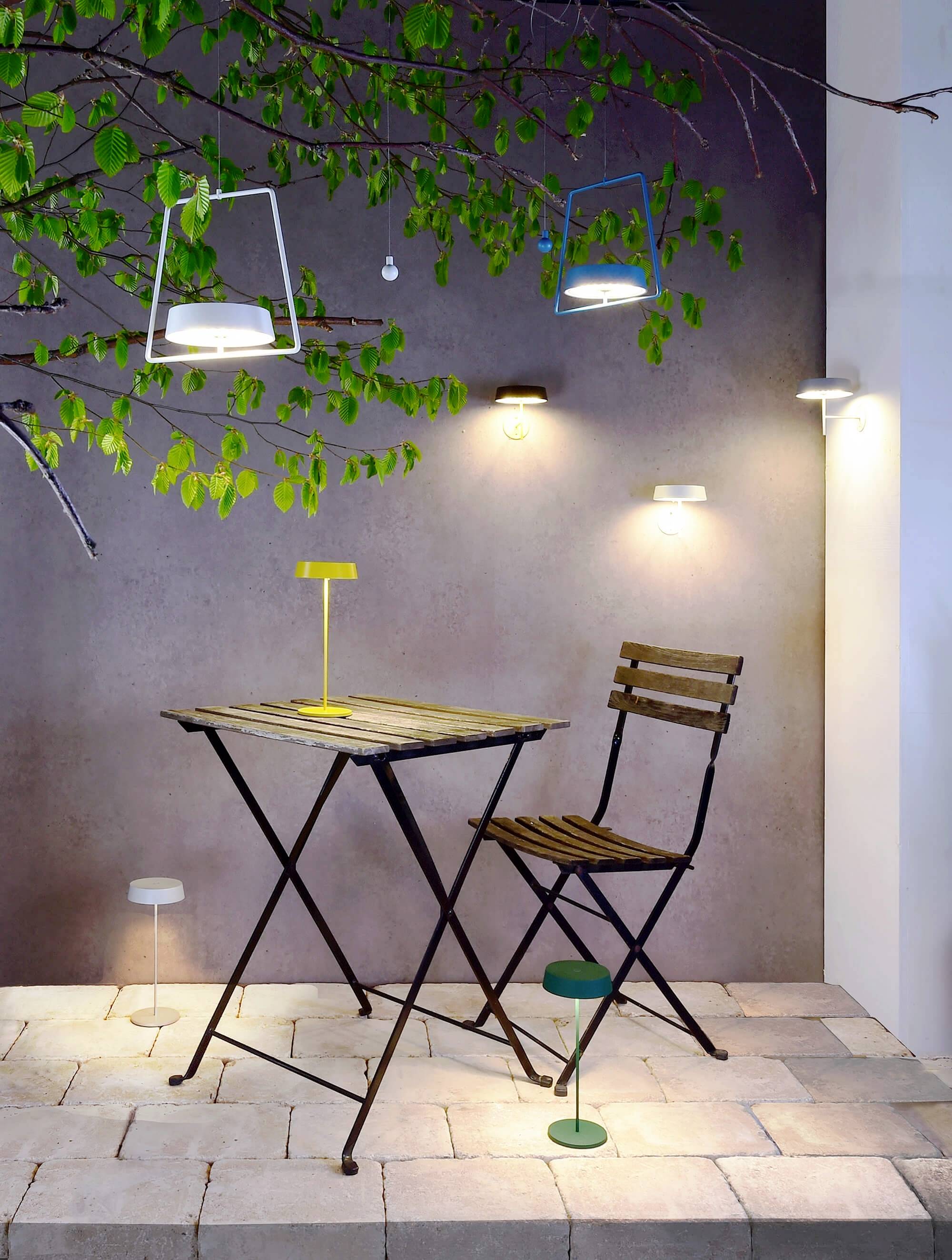 Záhrada s malým stolíkom a stoličkou, obklopená modernými lampami a zelenými listami, ktoré vytvárajú útulnú atmosféru.