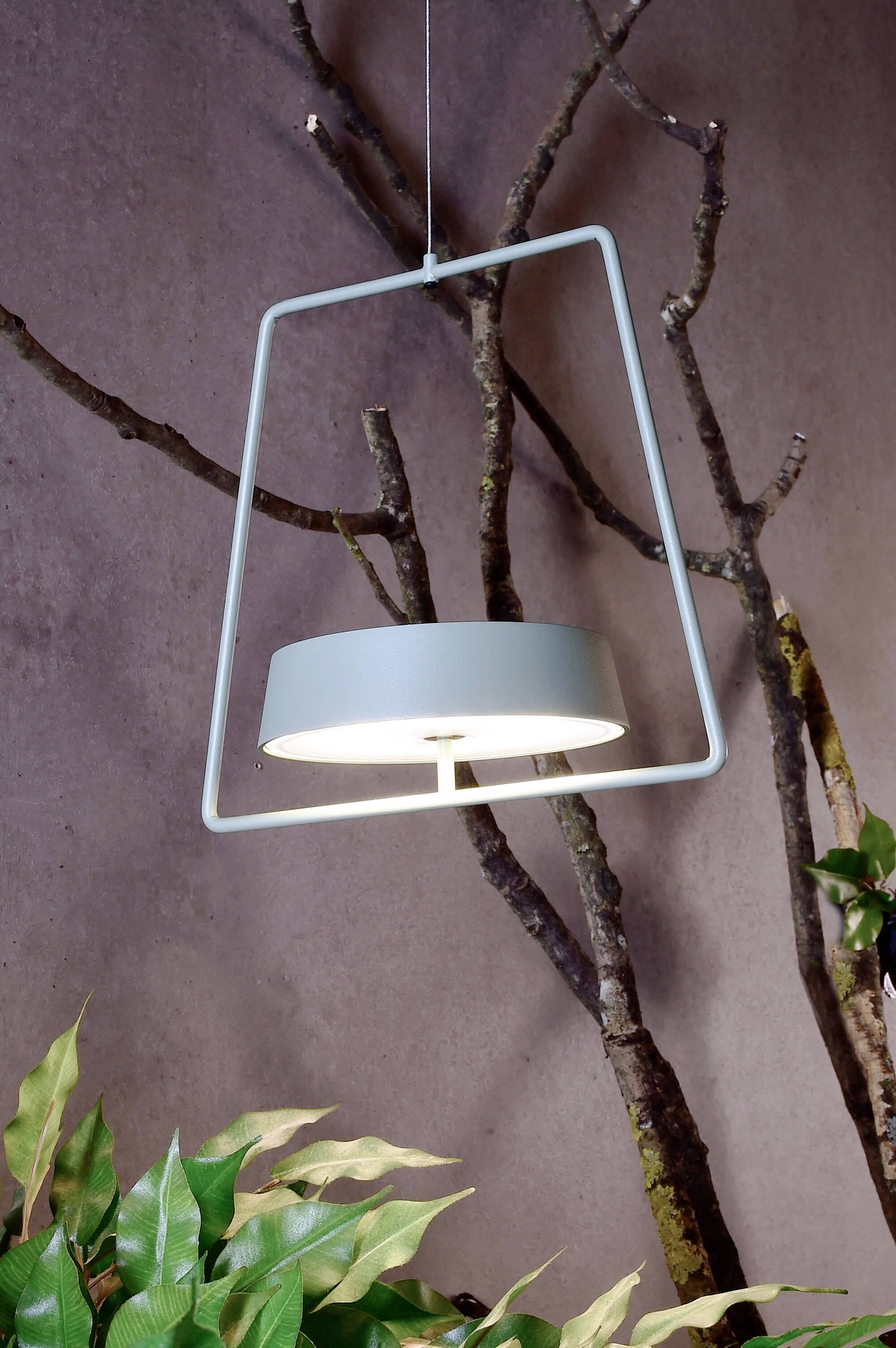 Moderná závesná lampa s obdĺžnikovým rámom pred sivou stenou s vetvami a listami, poskytuje štýlové osvetlenie.