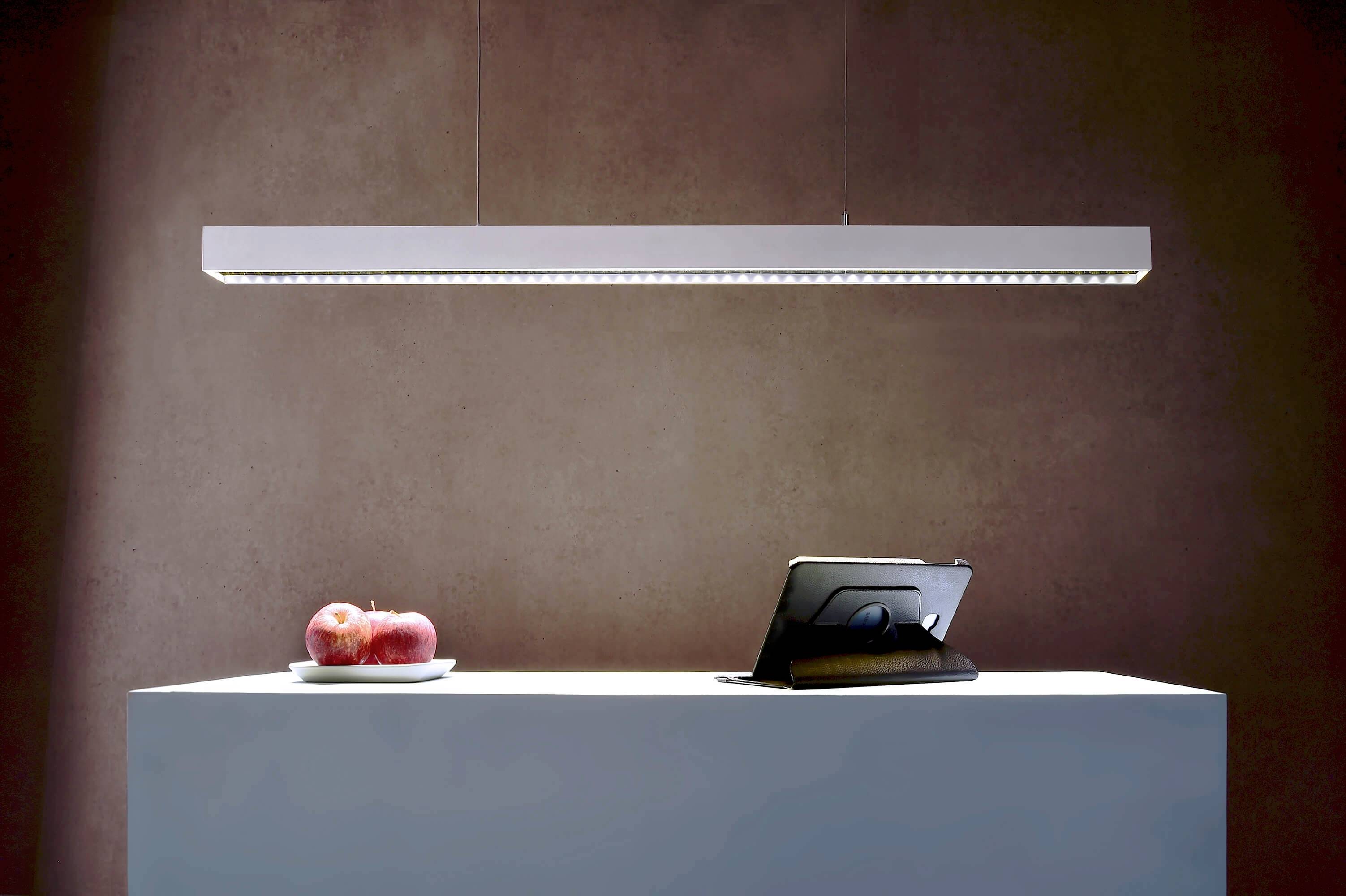 Moderný, obdĺžnikový závesný luster svieti nad stolom s tanierom červených jabĺk a stojaným tabletom.