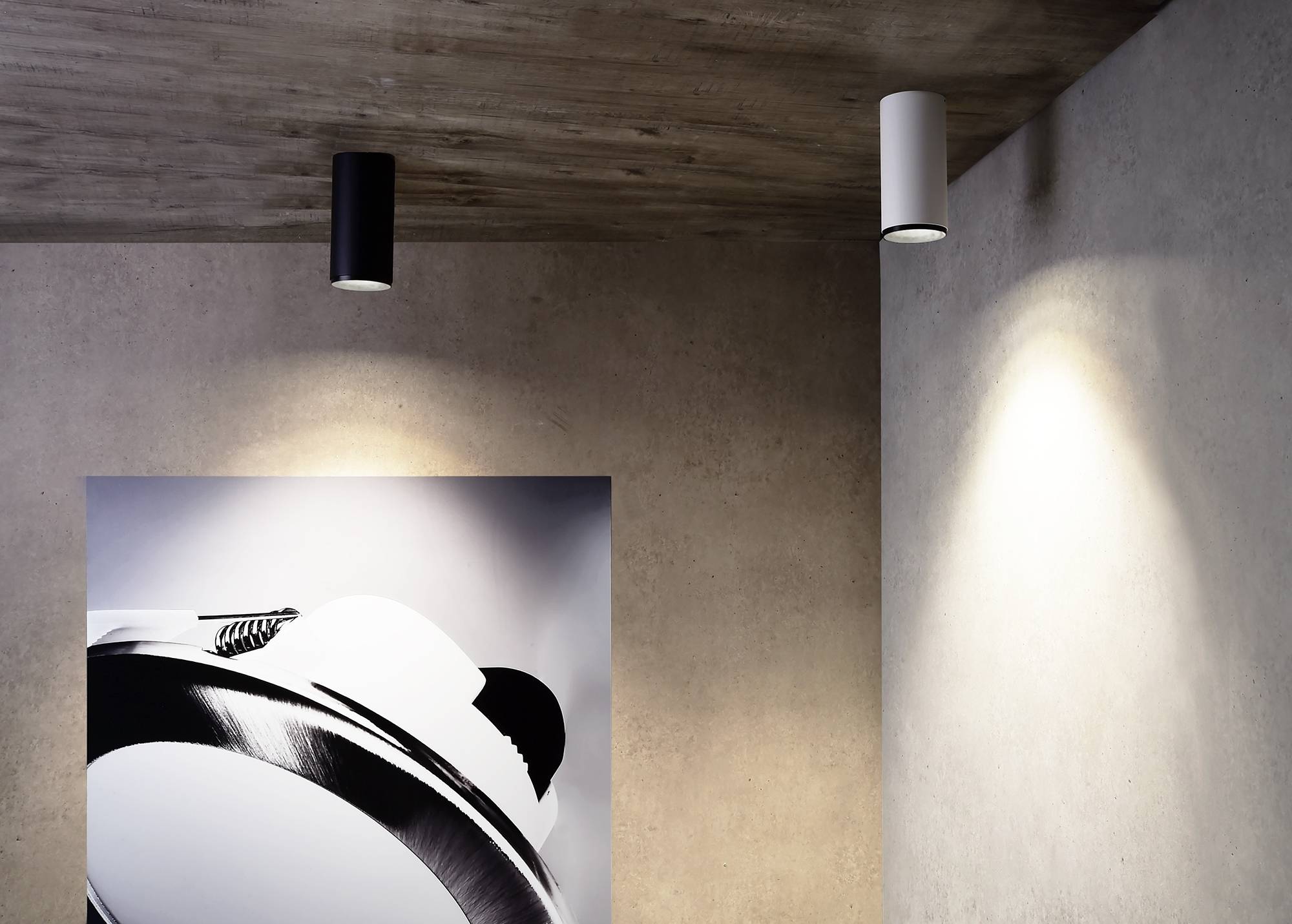 Minimalistický priestor s dreveným stropom, dvoma valcovými stropnými svietidlami a čierno-bielym umeleckým dielom na stene.