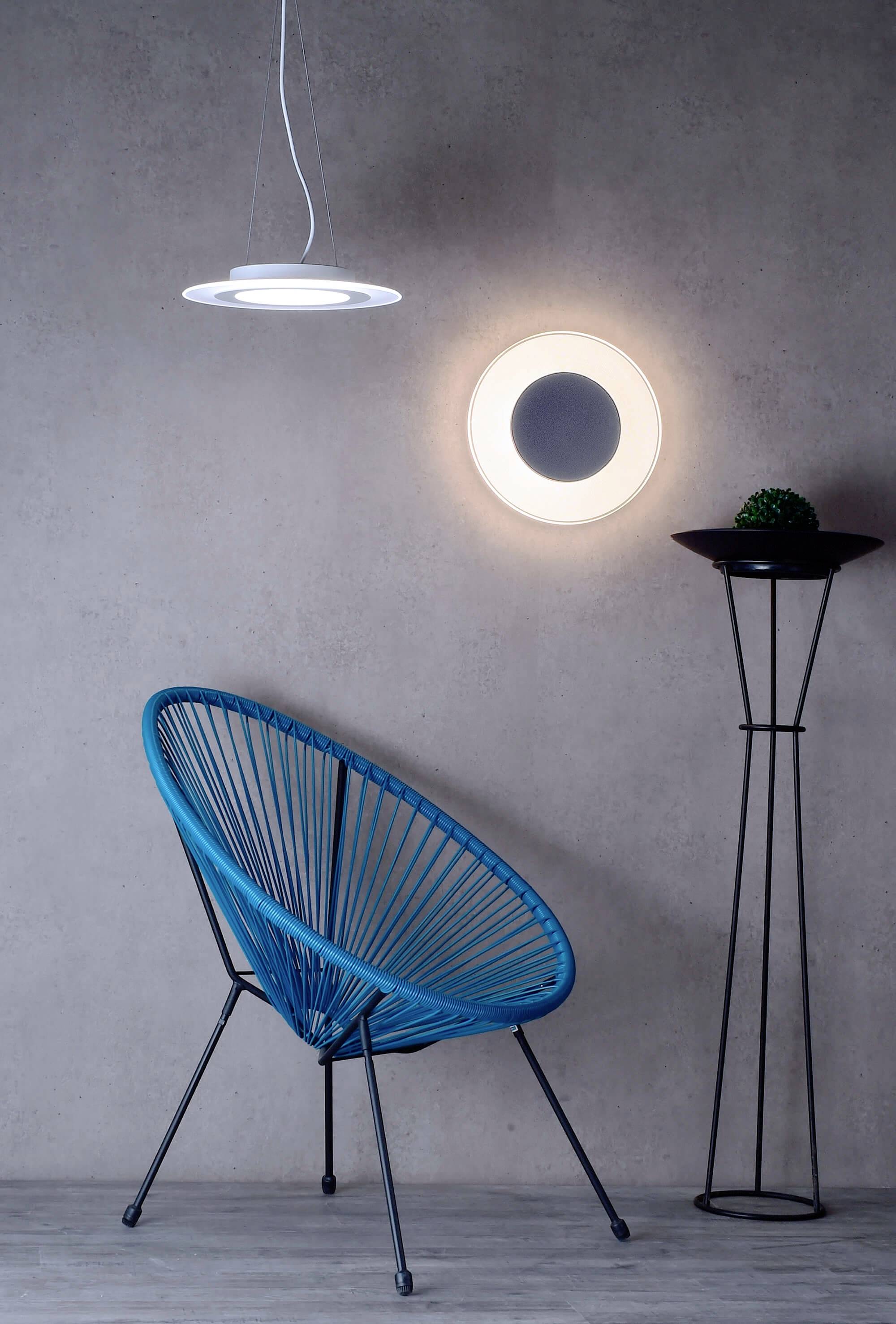 Minimalistický interiér s modrým stolíkom, moderným závesným svietidlom a okrúhlym, žiariacim zrkadlom na stene.