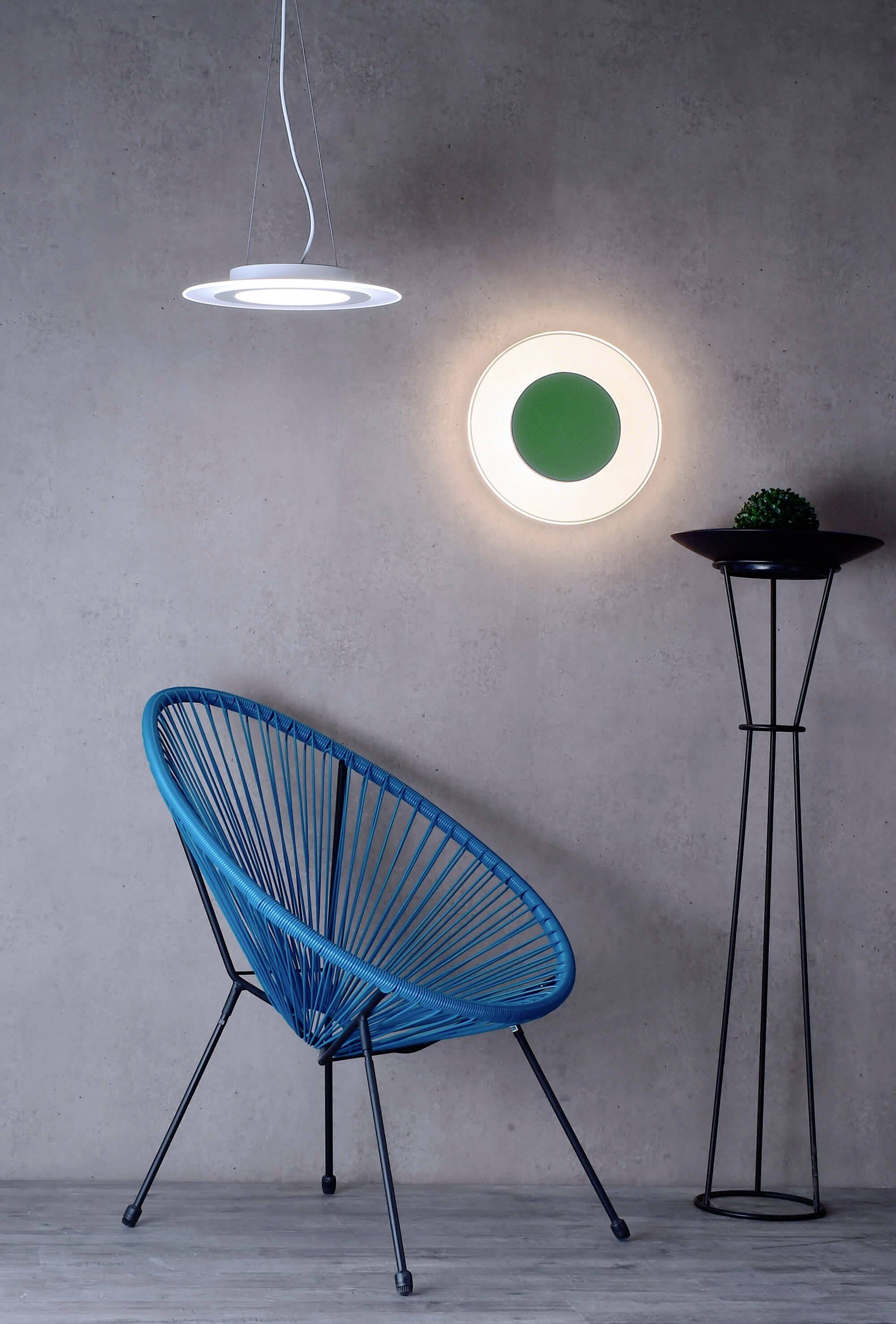 Modrá stolička s okrúhlym dizajnom stojí na tmavosivej podlahe pred sivou stenou. Vedľa nej svieti okrúhle nástenné svietidlo.