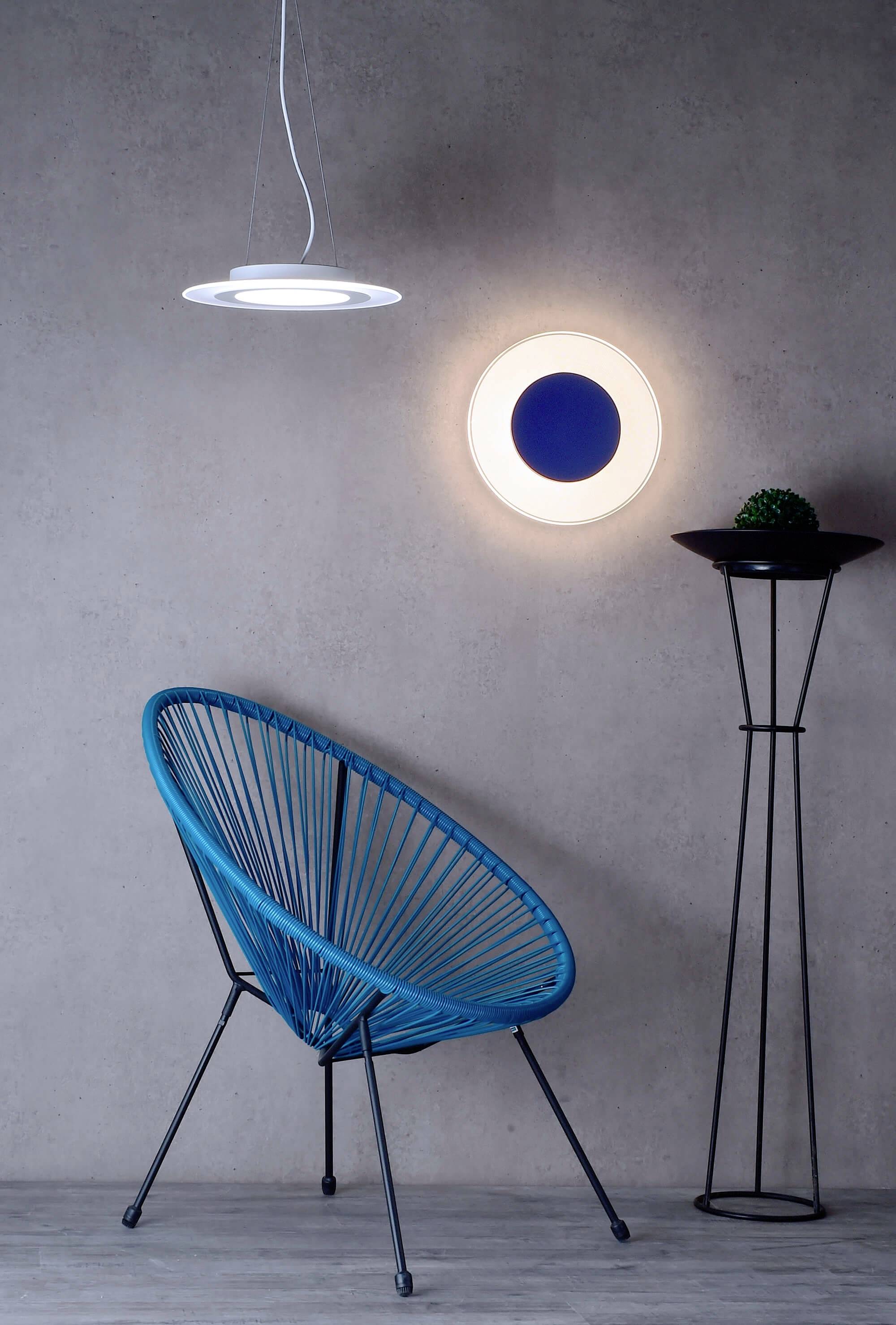 Modrá stolička s trojuholníkovým dizajnom stojí pred šedou stenou. Vpravo je na stene pripevnená moderná lampa.