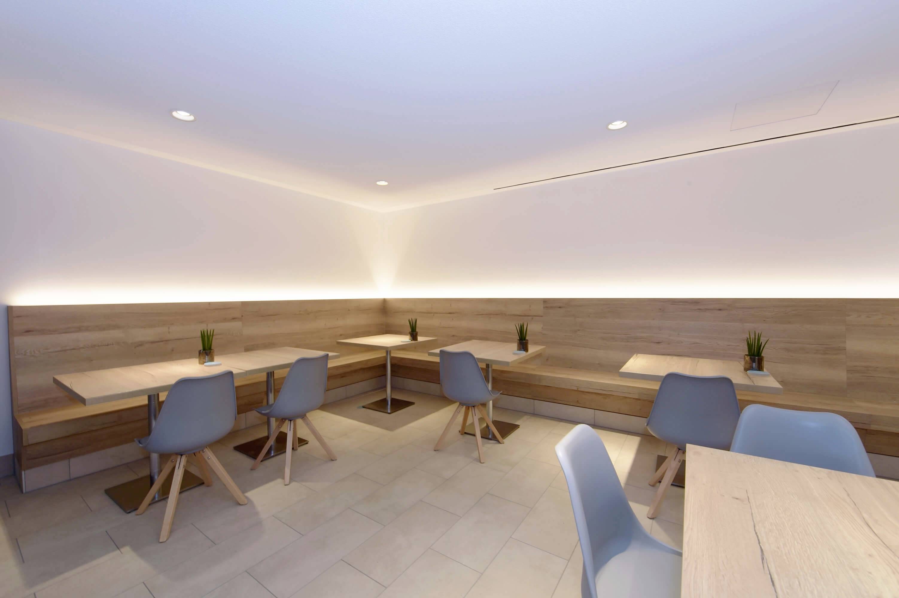 Moderná, prázdna jedáleň s drevenými stolmi, modrými stoličkami a malými rastlinami v rohovej časti s jemným osvetlením.