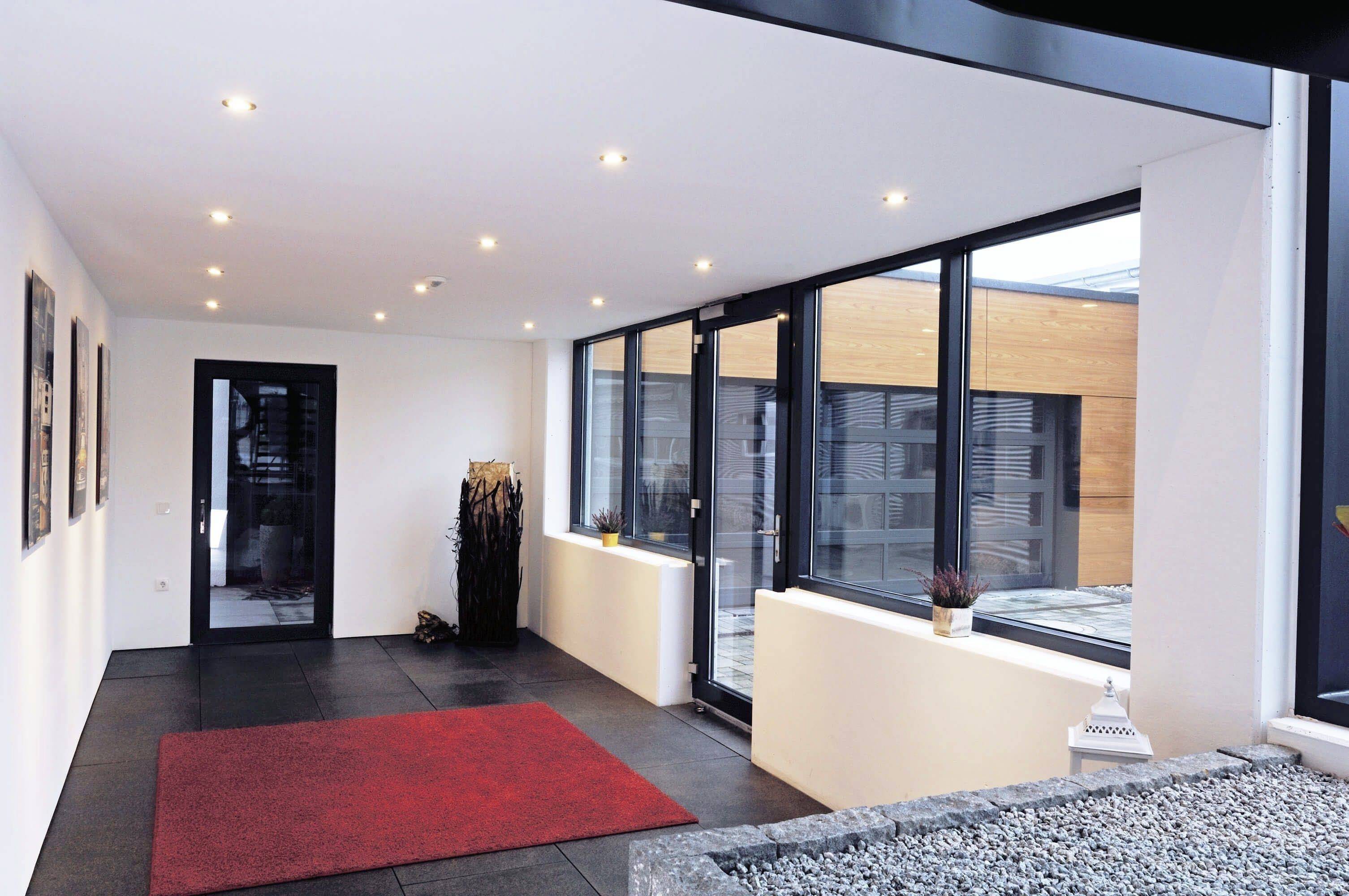 Moderná chodba s veľkými oknami, žiarivo červeným kobercom a rastlinami; spája vnútorný a vonkajší priestor, poskytuje veľa denného svetla.
