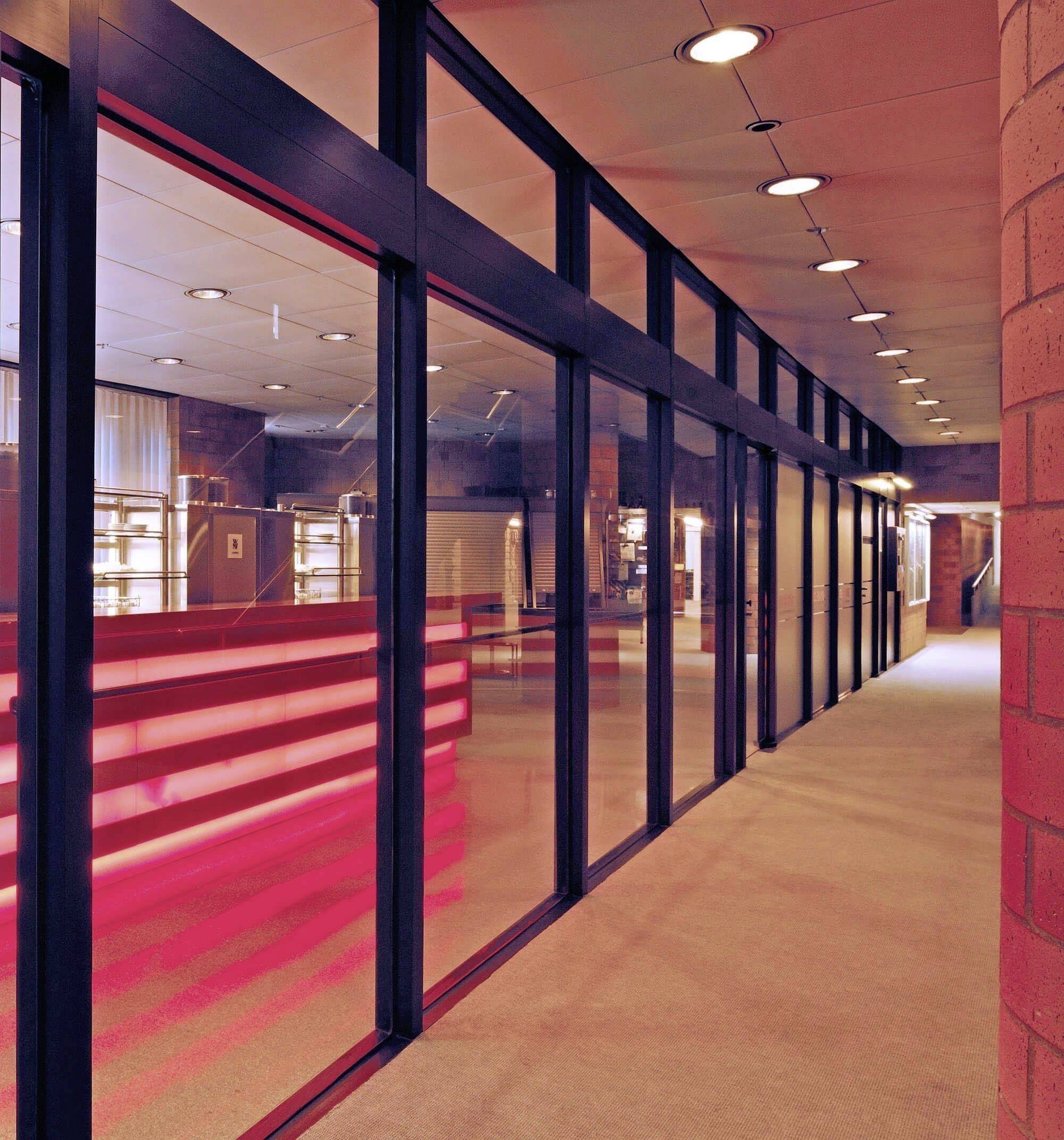 Chodba s modernou sklenenou stenou a štruktúrovanými stropnými svetlami; odráža teplé, naružovelé svetlo, ktoré vytvára štýlovú atmosféru.