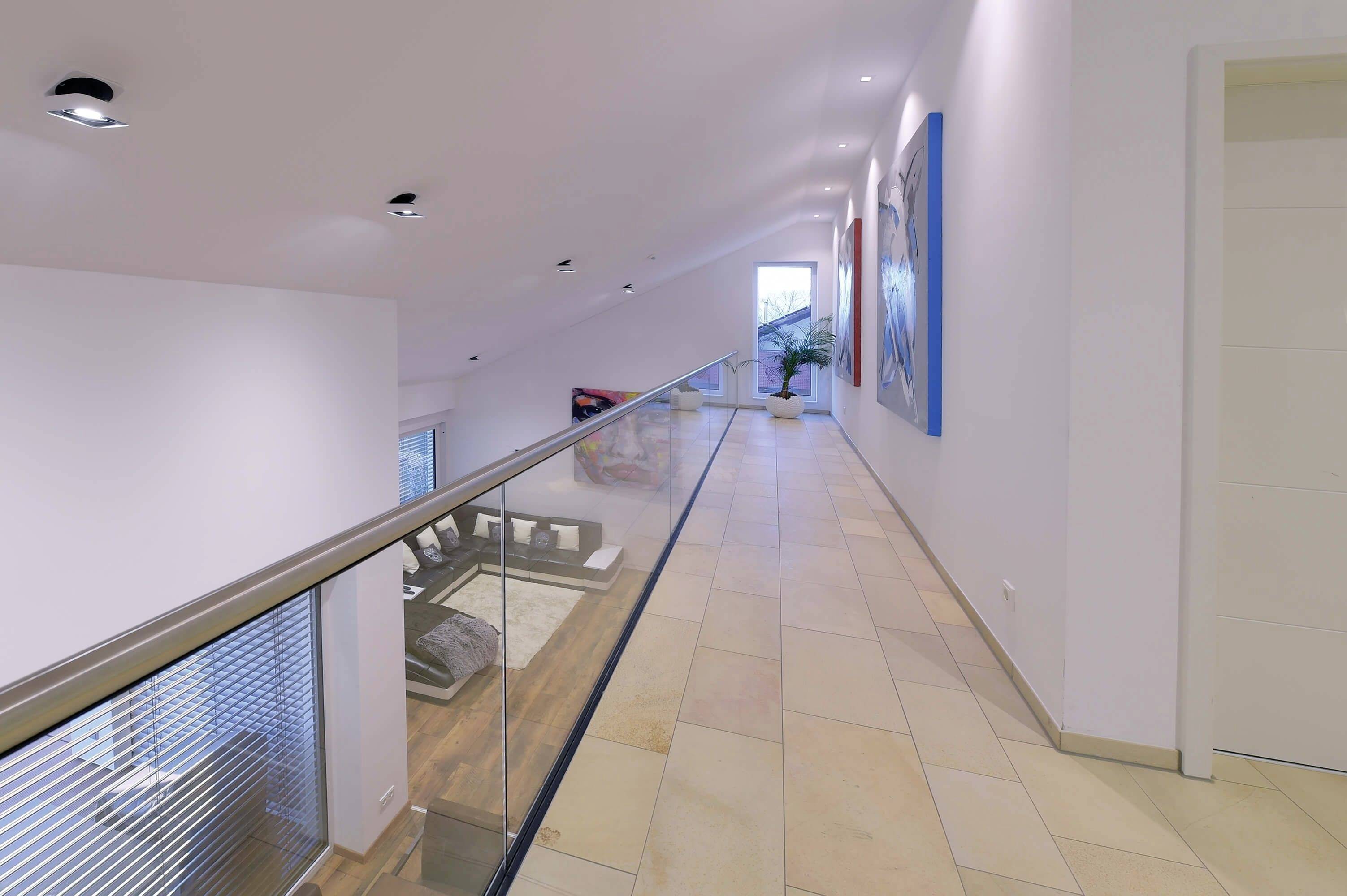 Moderná chodba so skleneným zábradlím, dlažbou a umením na stene, ktorá vedie k presklenej stene s výhľadom na svetlý priestor.