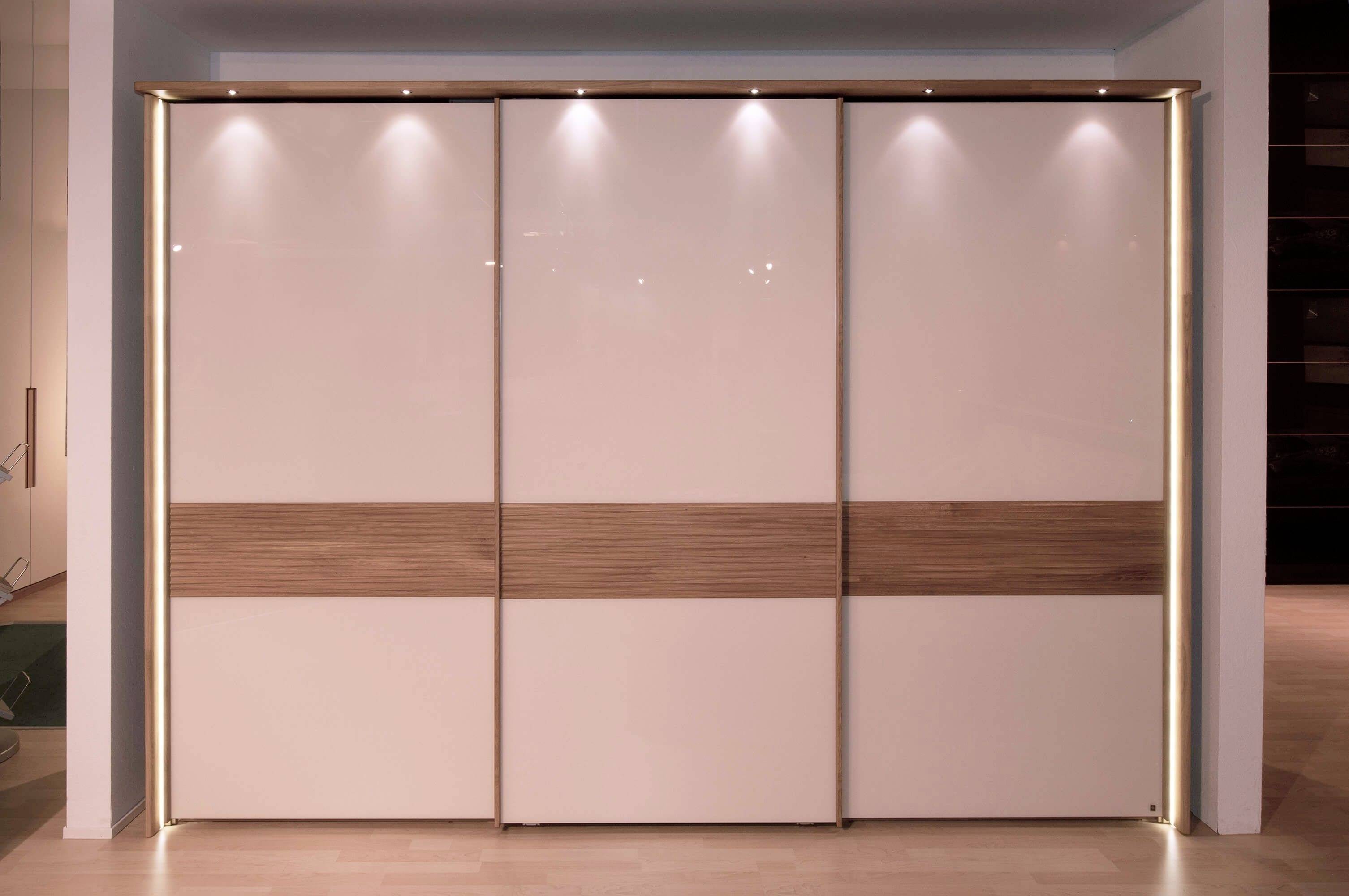 Moderná šatníková skriňa so štyrmi posuvnými dverami vyrobenými z lesklého materiálu s drevenými detailmi; úsporné LED svietidlá v hornej časti.