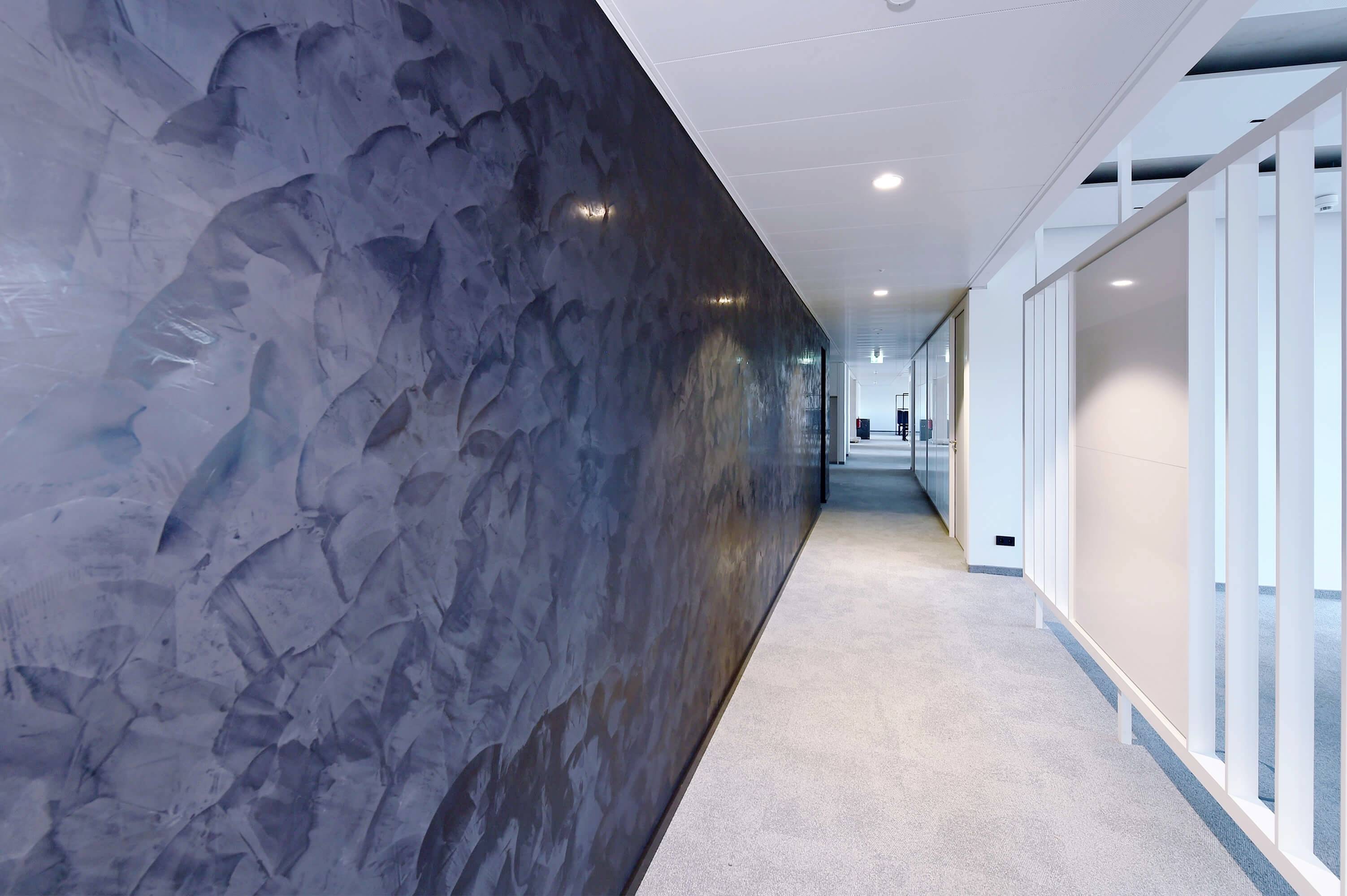 Dlhá, moderná chodba so tmavou stenou na ľavej strane a svetlým kobercom na podlahe. Na konci chodby sú viditeľné kancelárske dvere.