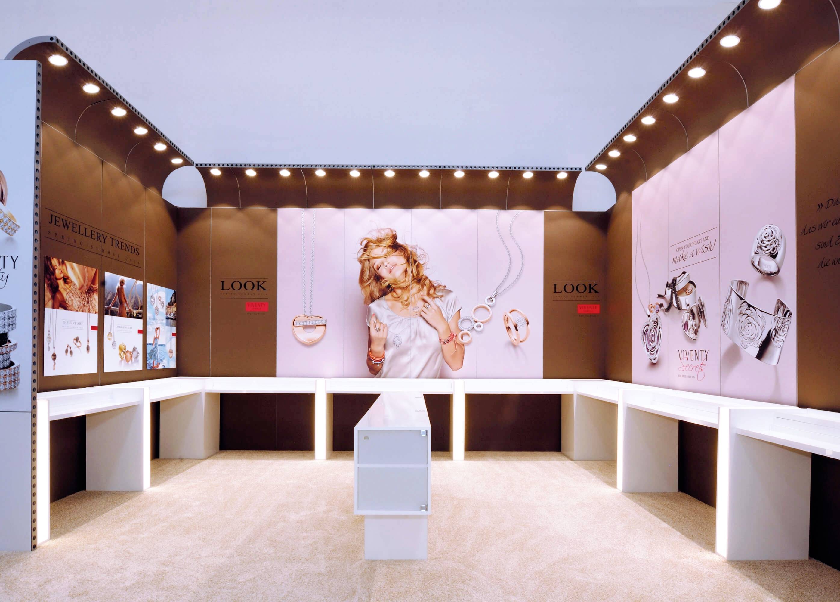 Predajný priestor s prezentáciami šperkov na hnedých stenách, osvetlený stropnými reflektormi. V strede biely stôl.