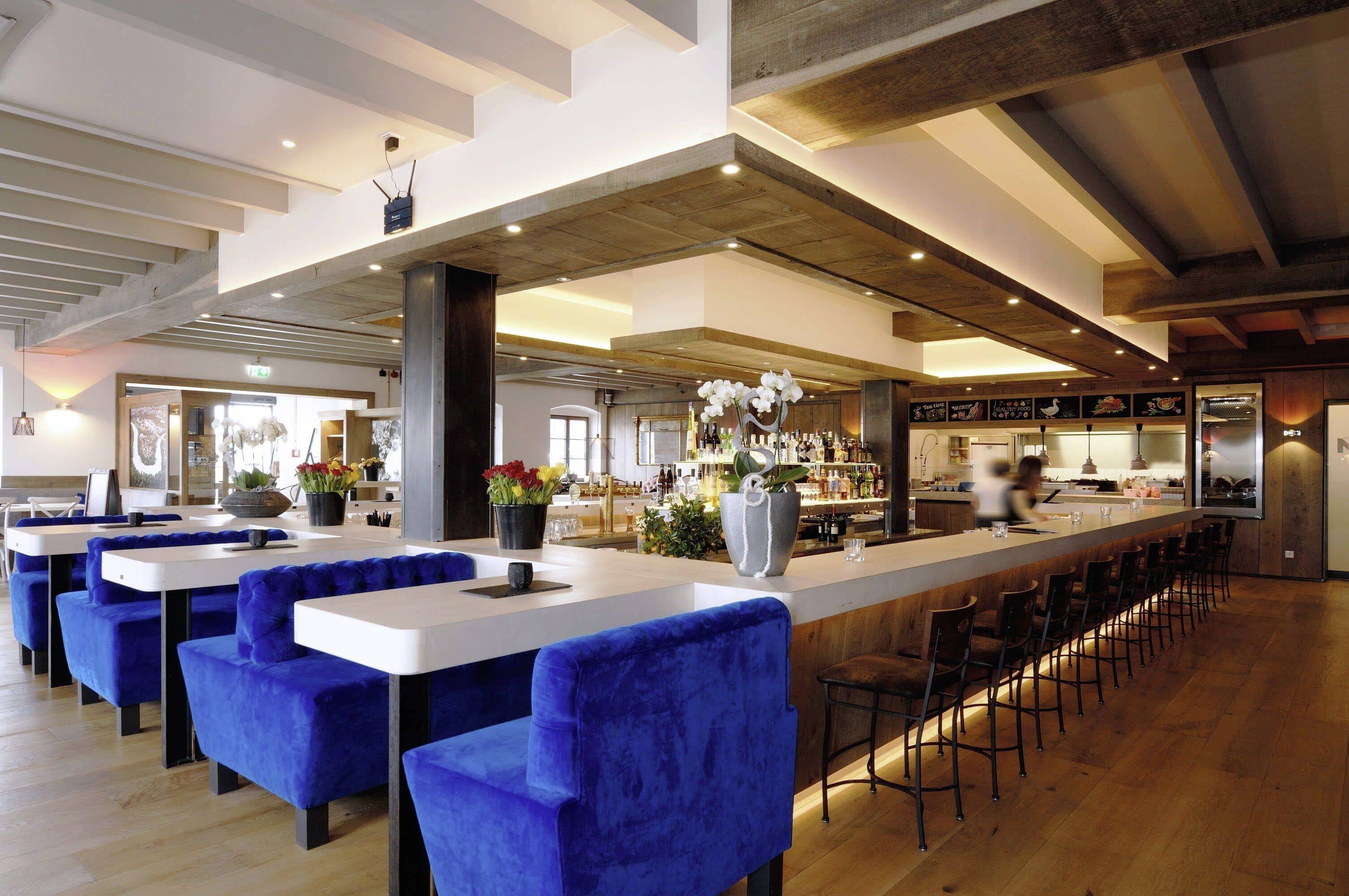 Moderná reštaurácia so svetlomodrými čalúnenými stoličkami, drevenou podlahou, dlhým barovým pultom a atmosférickým osvetlením.