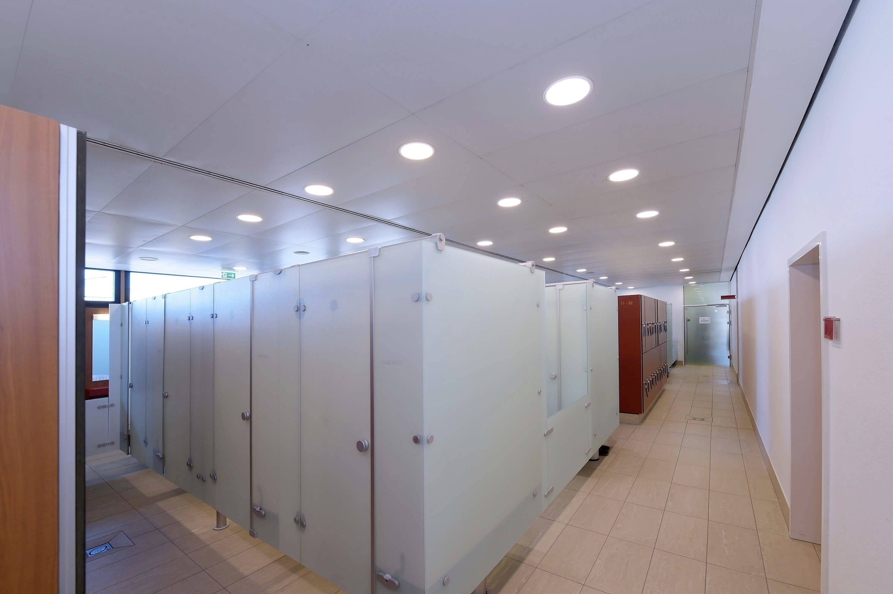 Veľká, čistá umyváreň s niekoľkými presklenými sprchovými kabínkami na ľavej strane. Svetlý priestor s dlaždicovými podlahami a stenami.